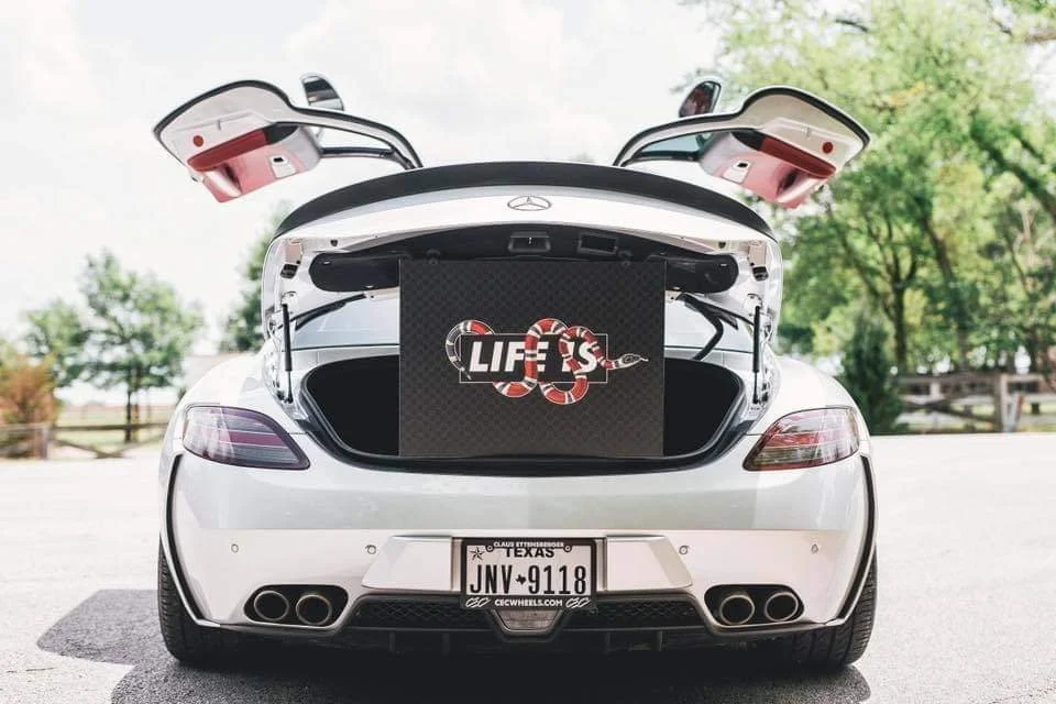 White Mercedes-Benz with open trunk and gullwing doors, parked outdoors with green trees in the background, displaying a LIFE'S logo with a snake.