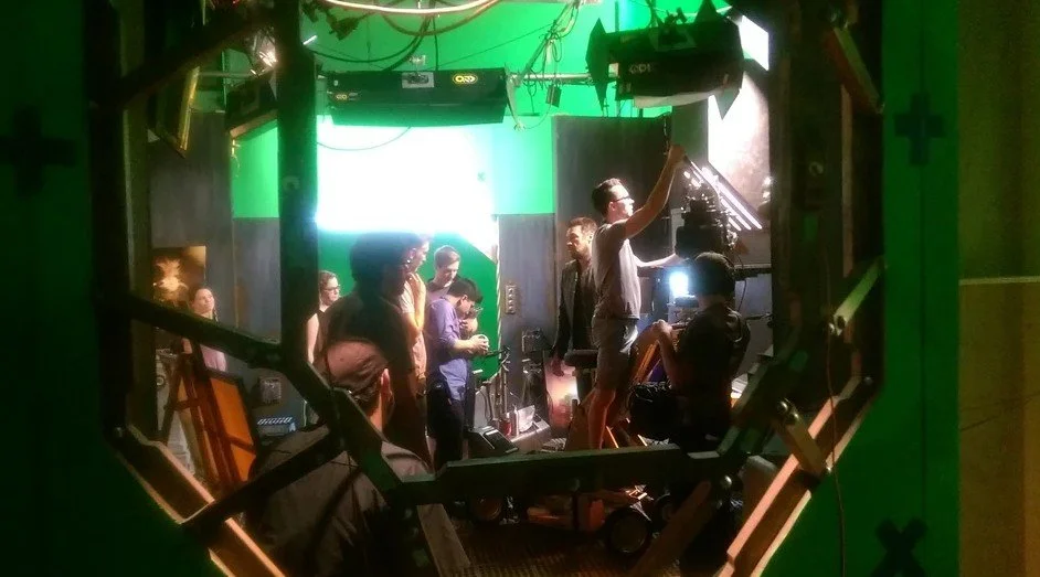 Behind-the-scenes view of a film or TV production crew working with a green screen as the backdrop, with multiple crew members operating equipment and preparing for a shoot.