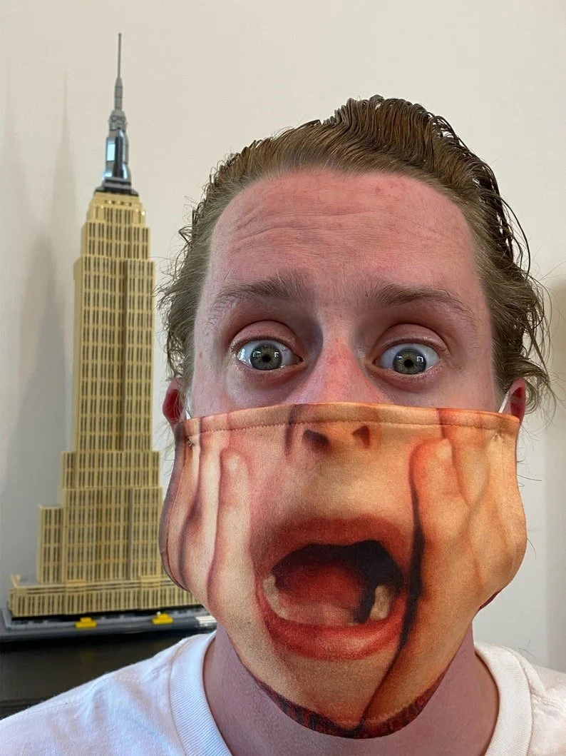 Person wearing a mask with a large mouth and teeth design, wide eyes, and a backdrop of a model of the Empire State Building.