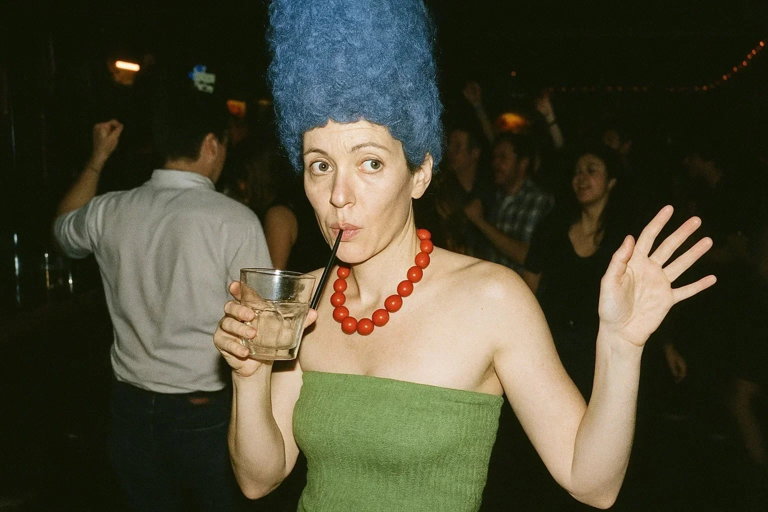A woman with a tall blue wig, straw hat, and red beads on her neck drinks from a glass with a straw at a party, wearing a green strapless dress.
