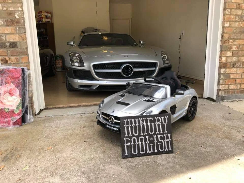 A garage with a silver Bugatti car inside and a small, silver toy Mercedes-Benz car outside with a sign that reads 'HUNGRY FOOLISH' placed in front of it.