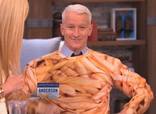A man on a TV show wearing a shirt with a design resembling French fries, sitting and smiling at a woman with blonde hair who is partially visible.