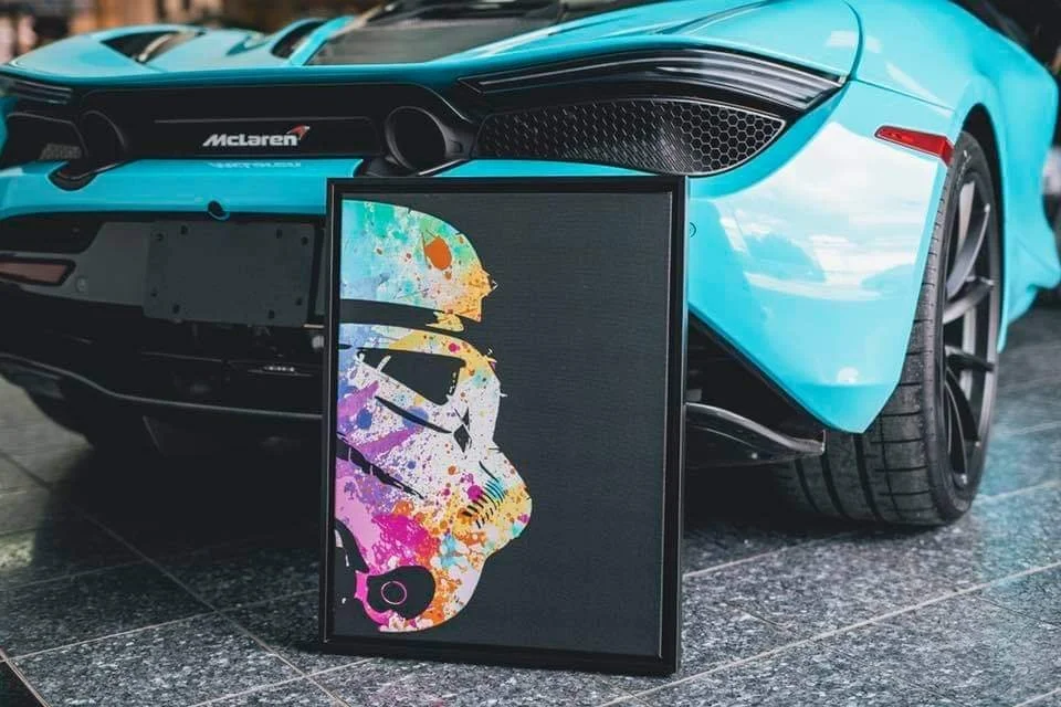A turquoise McLaren sports car with black accents and large tires, parked on a tiled surface, with a colorful framed art print of a stormtrooper from Star Wars leaning against the car.