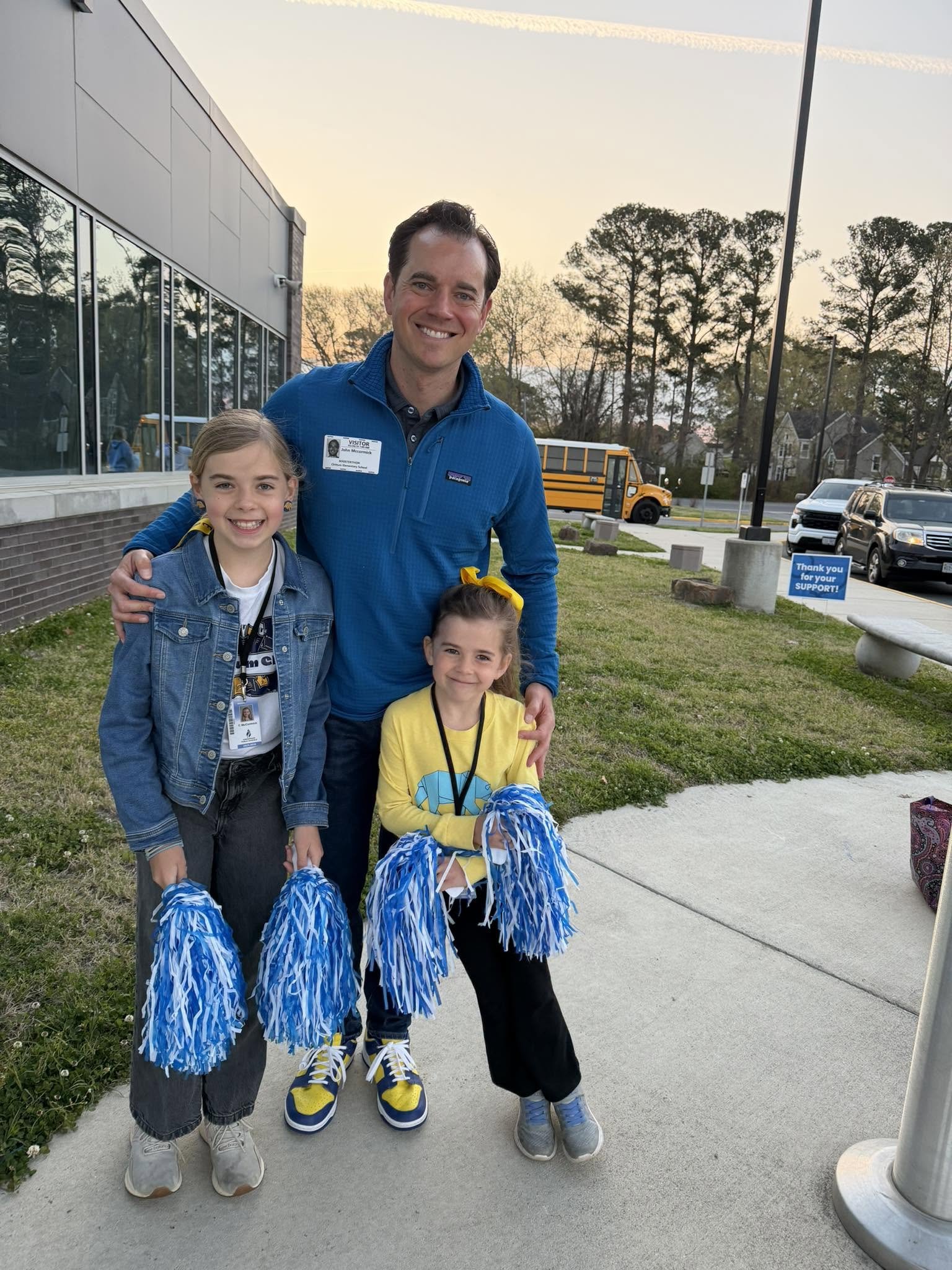 A group of three children and an adult man standing outside, smiling and holding blue and white pom-poms, in front of a building and a parking lot at sunset.