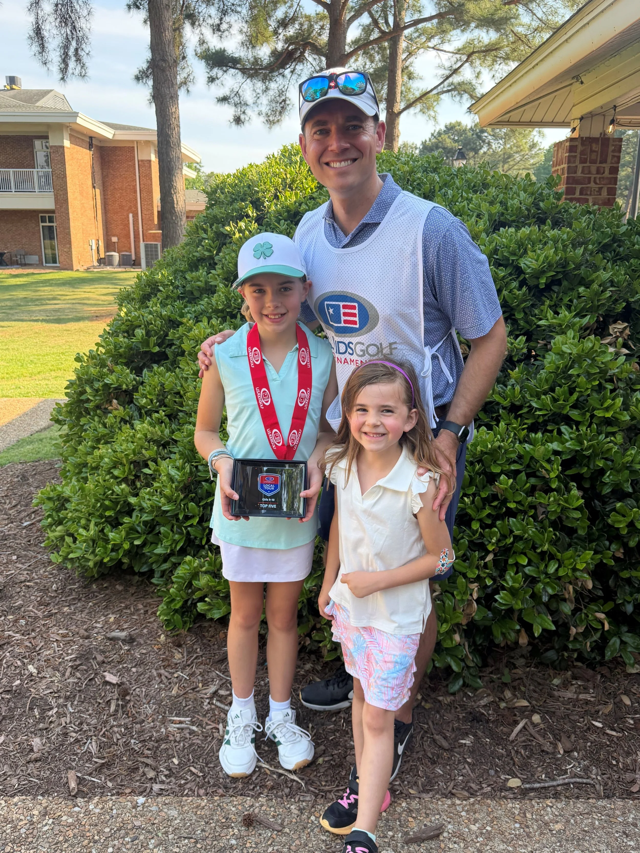 A group of three children and one adult male standing outdoors in a garden area with bushes and trees, with buildings in the background. The children are smiling and one is holding a plaque. The adult is wearing a sports shirt and the children are dressed casually.