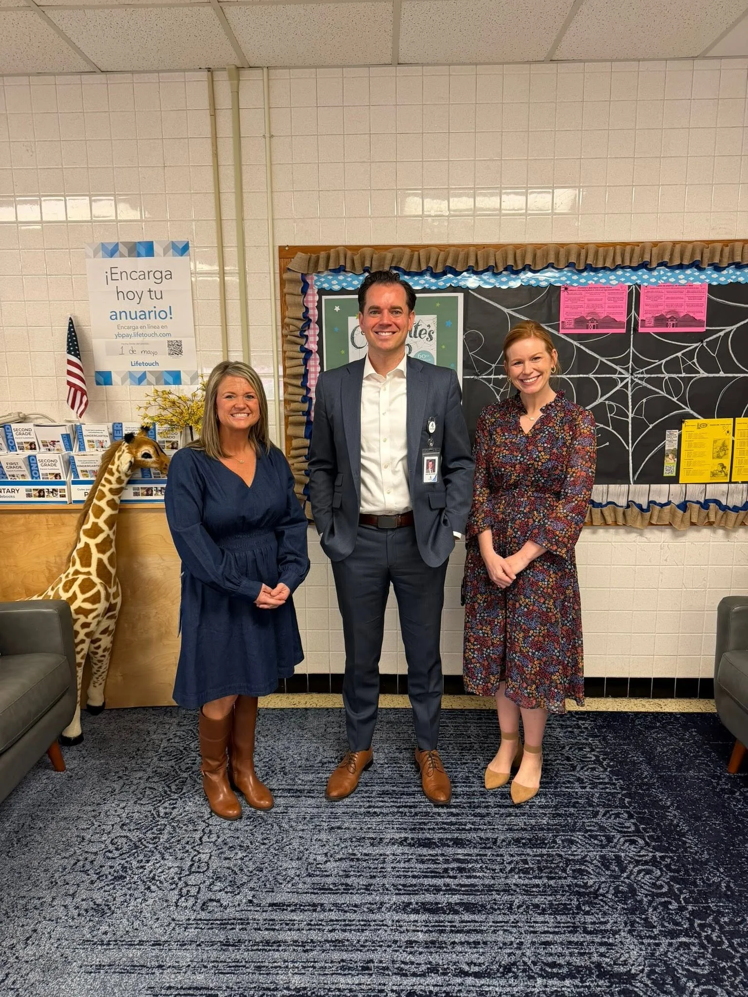 Three people are standing and smiling in front of a decorated blackboard with web webs. The woman on the left is wearing a dark blue dress and brown boots, with a stuffed giraffe toy beside her. The man in the middle is wearing a suit with an ID badge, and the woman on the right is wearing a floral dress and yellow heels. There is an American flag, yellow flowers, and flyers on a table in the background.