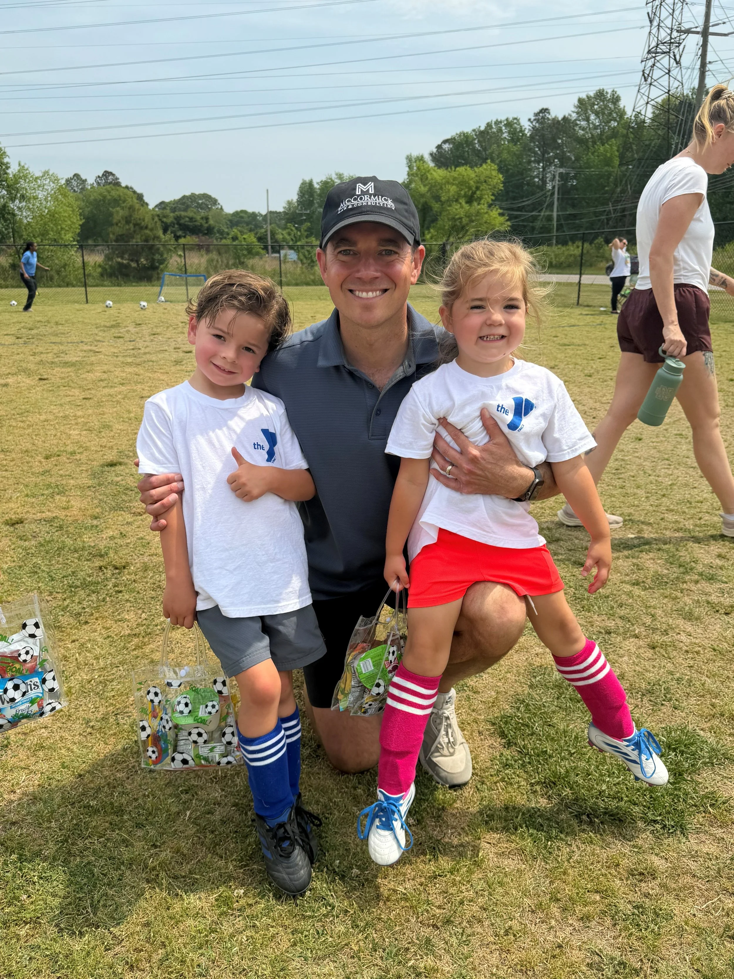 A man kneeling on the grass with two children, all smiling at the camera during a daytime outdoor soccer practice or event, with other children and soccer balls in the background.