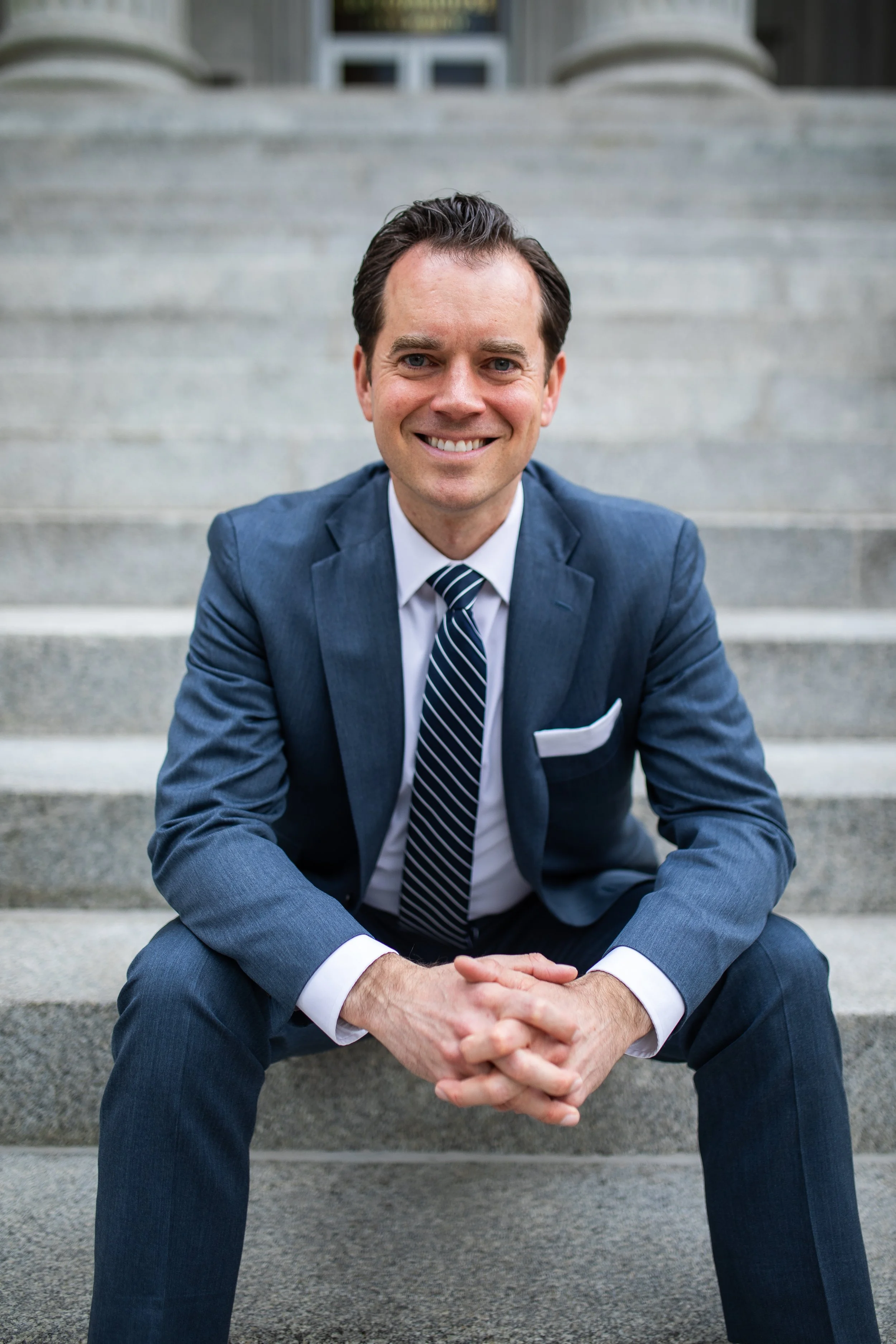 A man in a blue suit with a white shirt and striped tie sitting on outdoor concrete staircase, smiling at the camera.