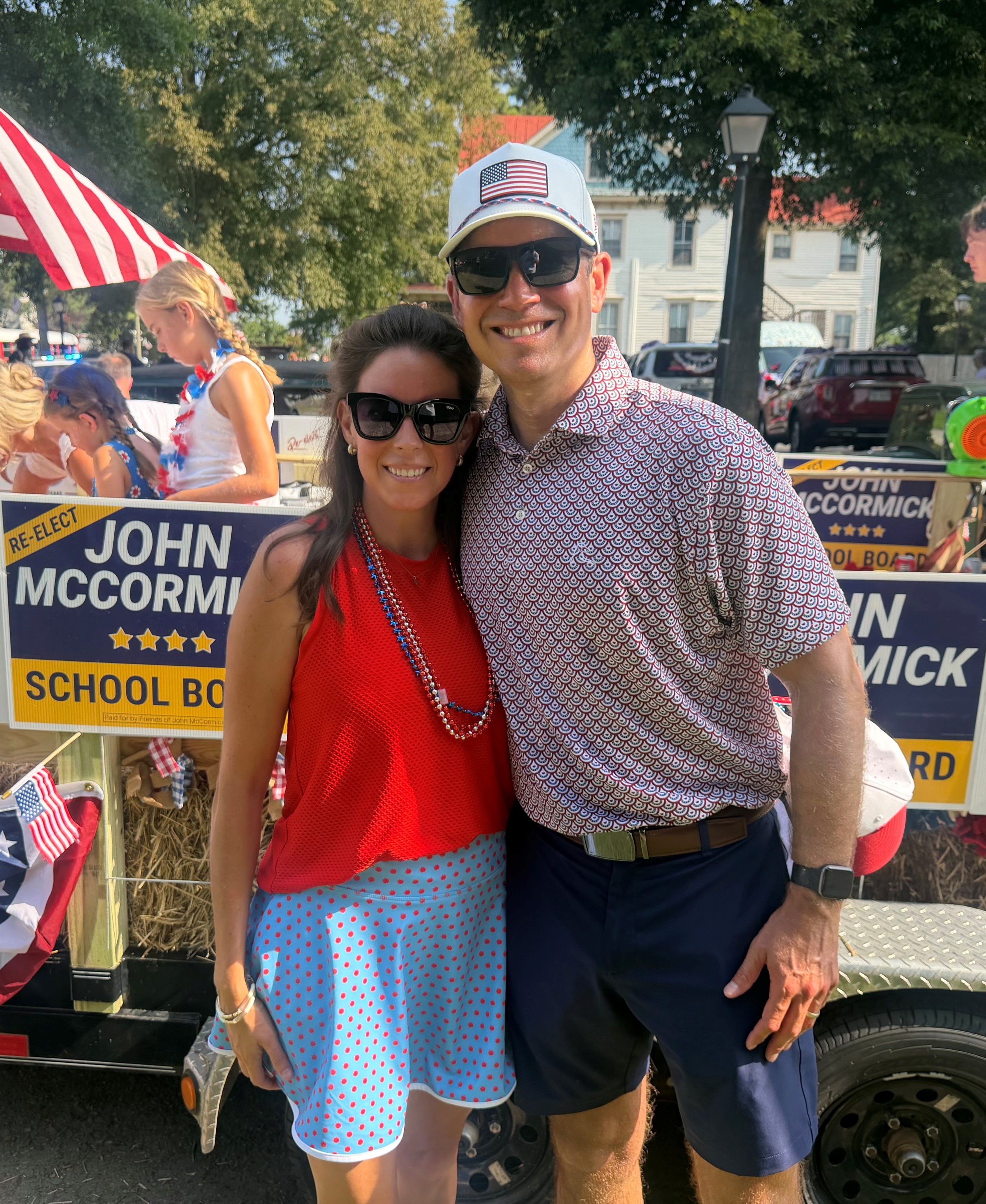 A man and woman smiling together outdoors at a patriotic event, standing in front of a parade float with political campaign signs for John McCormick. The woman wears a red top and a light blue polka dot skirt, while the man wears a patterned shirt, dark shorts, a white cap with an American flag patch, and sunglasses.