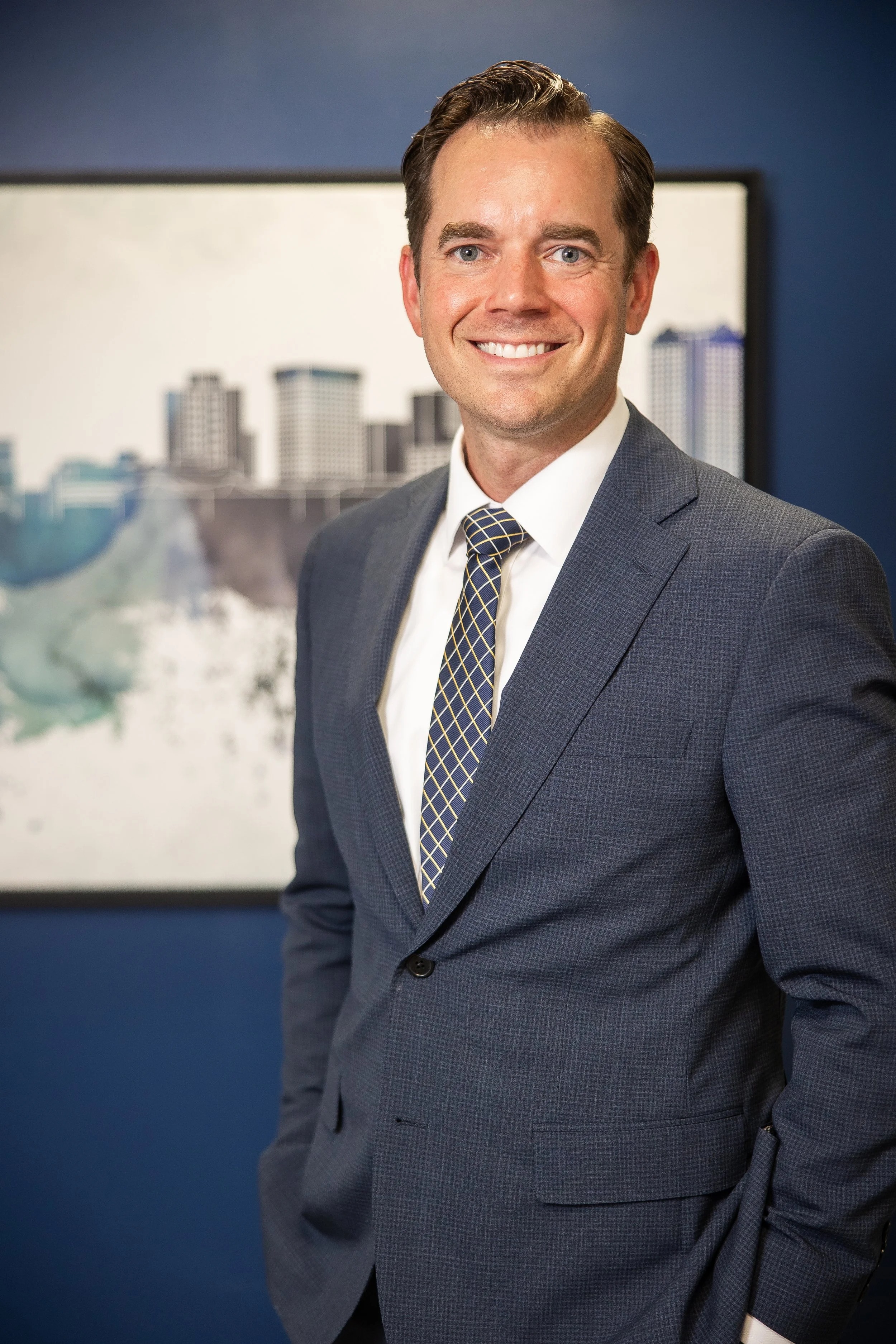 A man in a business suit standing in front of a cityscape painting, smiling at the camera.