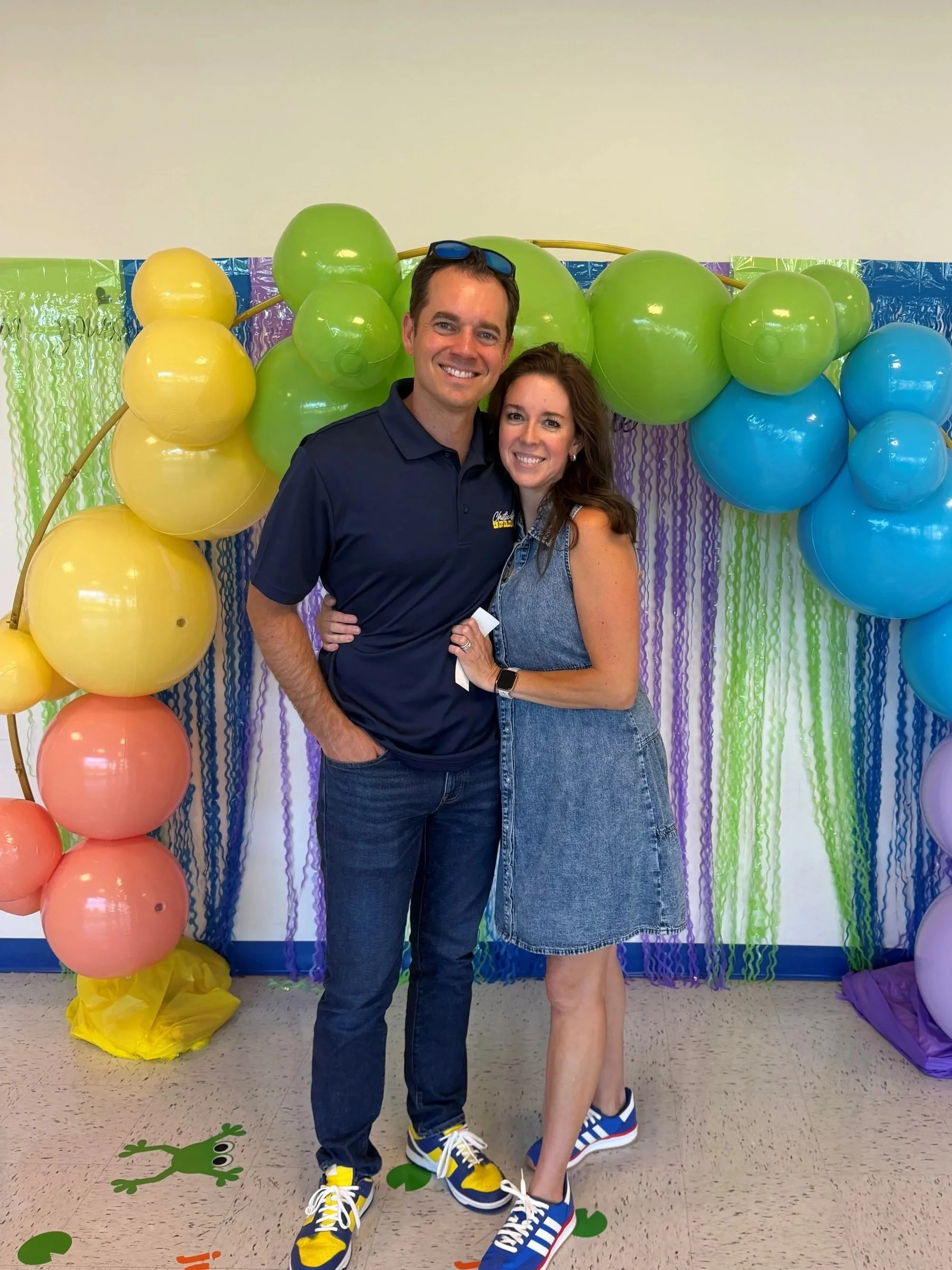 A man and woman smiling in front of a colorful balloon backdrop with purple, green, yellow, and blue balloons and curtain decorations on the wall.