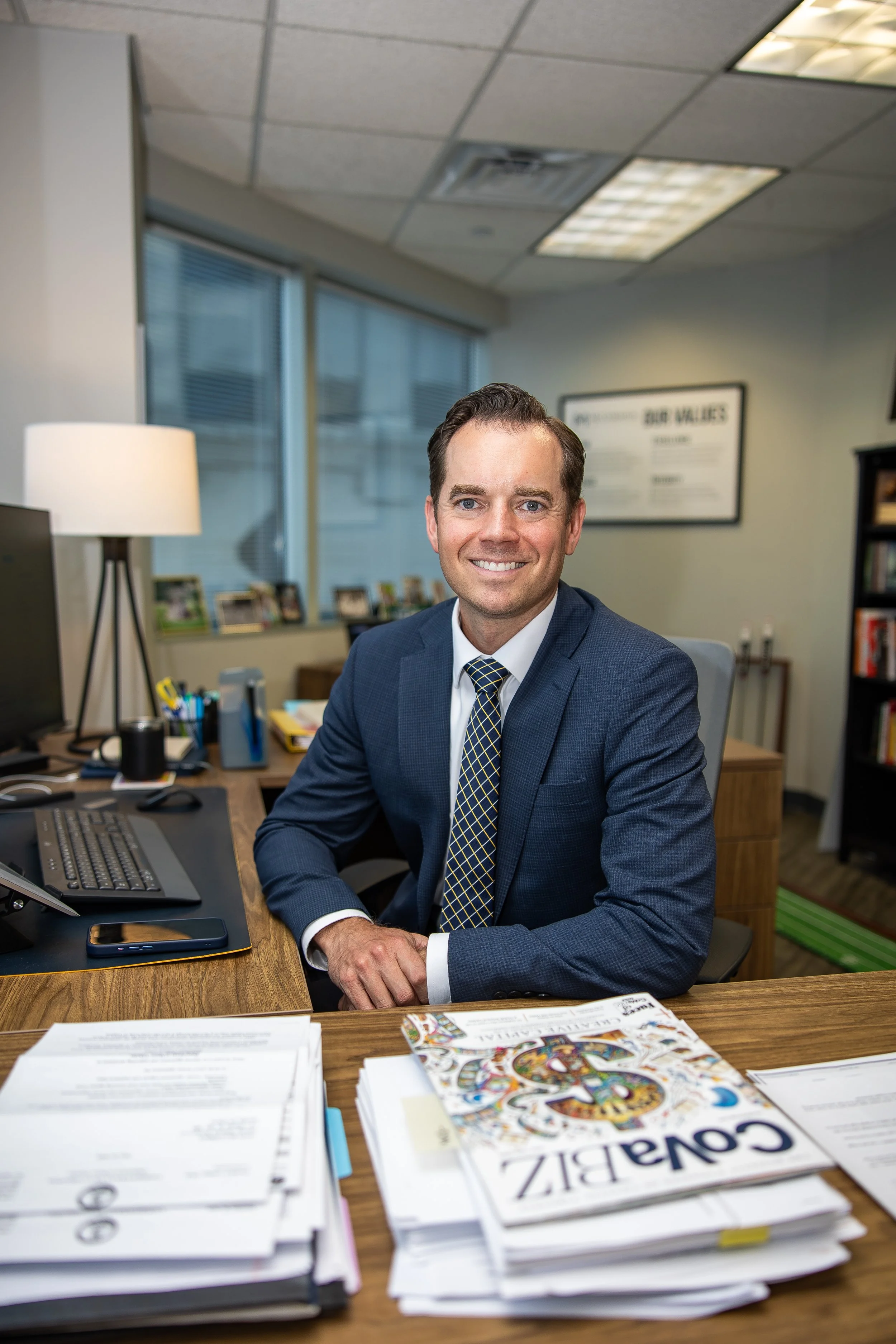 A man in a suit sitting at a cluttered office desk, smiling at the camera, with books, papers, and a computer.