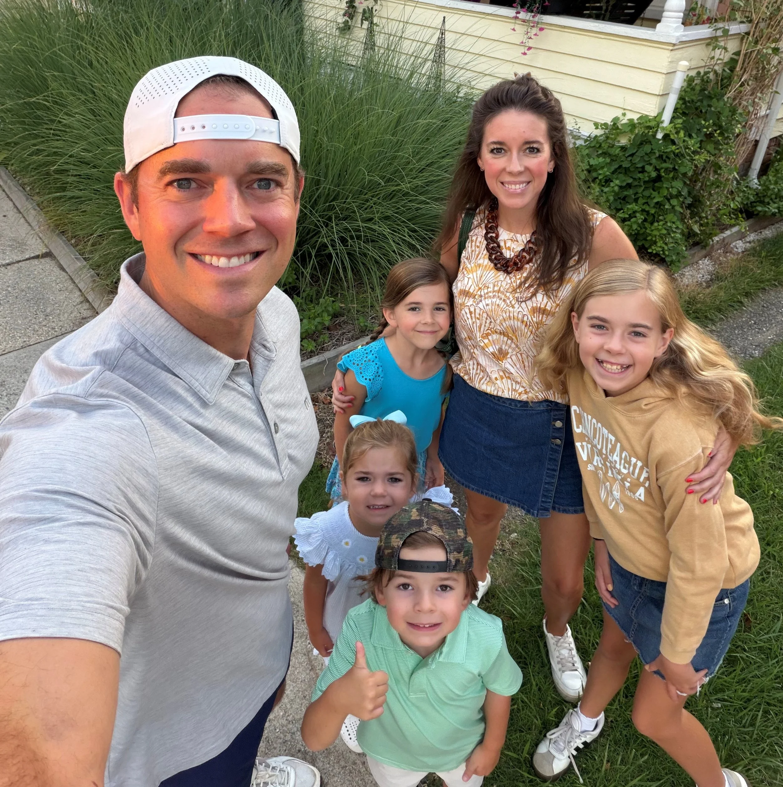 A group of children and adults smiling for a selfie outdoors with greenery and a yellow house in the background.