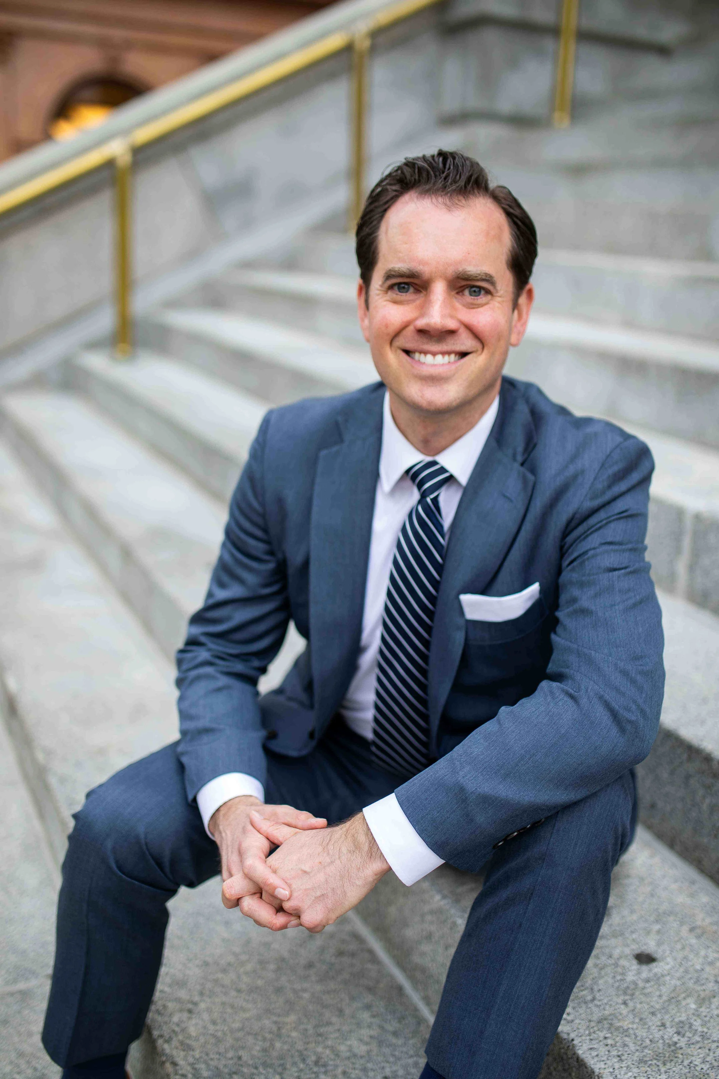 A man in a blue suit with a white shirt and striped tie, sitting on concrete stairs, smiling at the camera with his hands clasped.