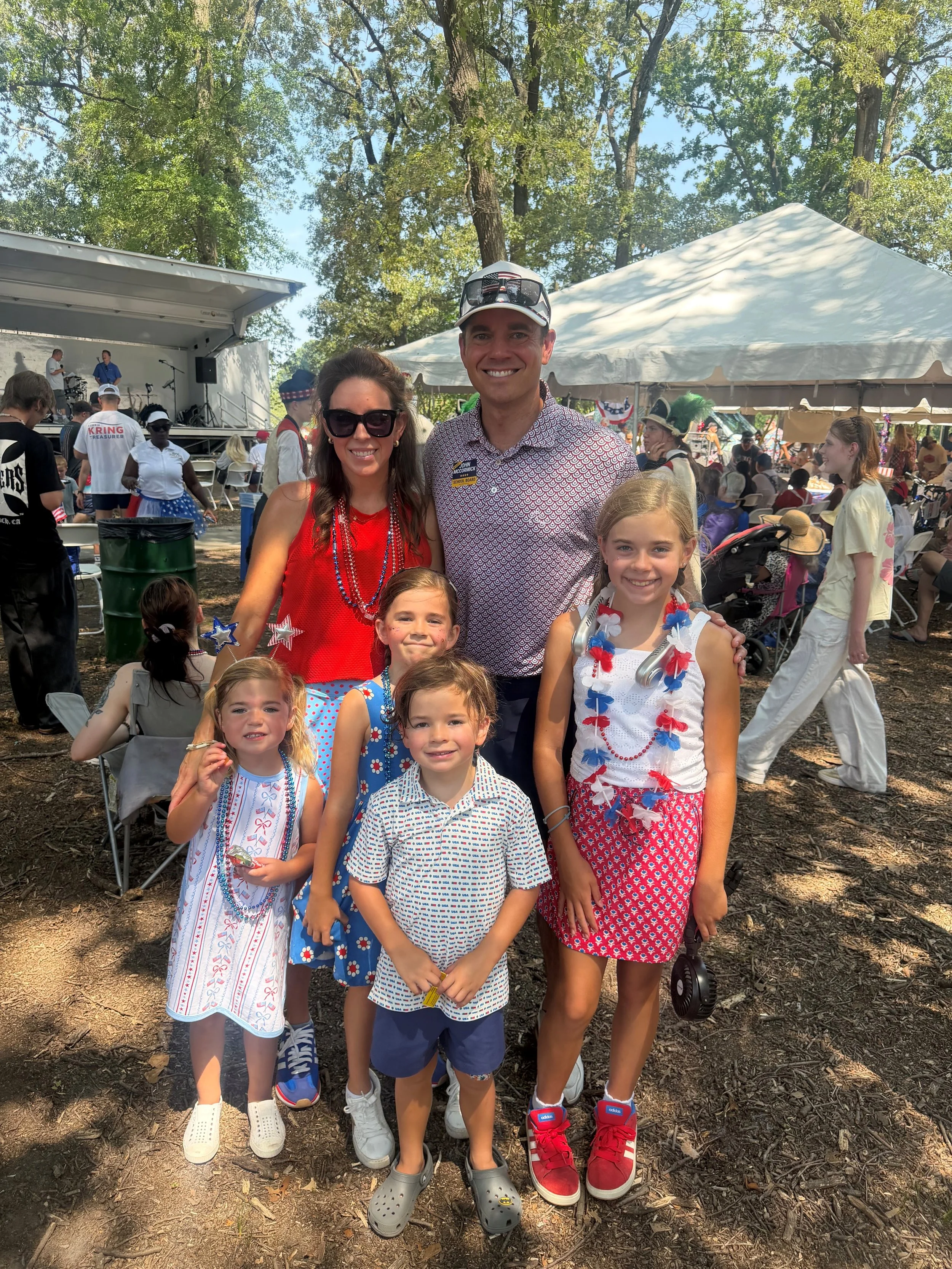 A group of children and adults celebrating at an outdoor event during the daytime. The children are dressed in patriotic red, white, and blue outfits with star and flag decorations. Two adults and three children are smiling and posing for the photo in front of a crowd, with a stage and a white tent visible in the background surrounded by trees.