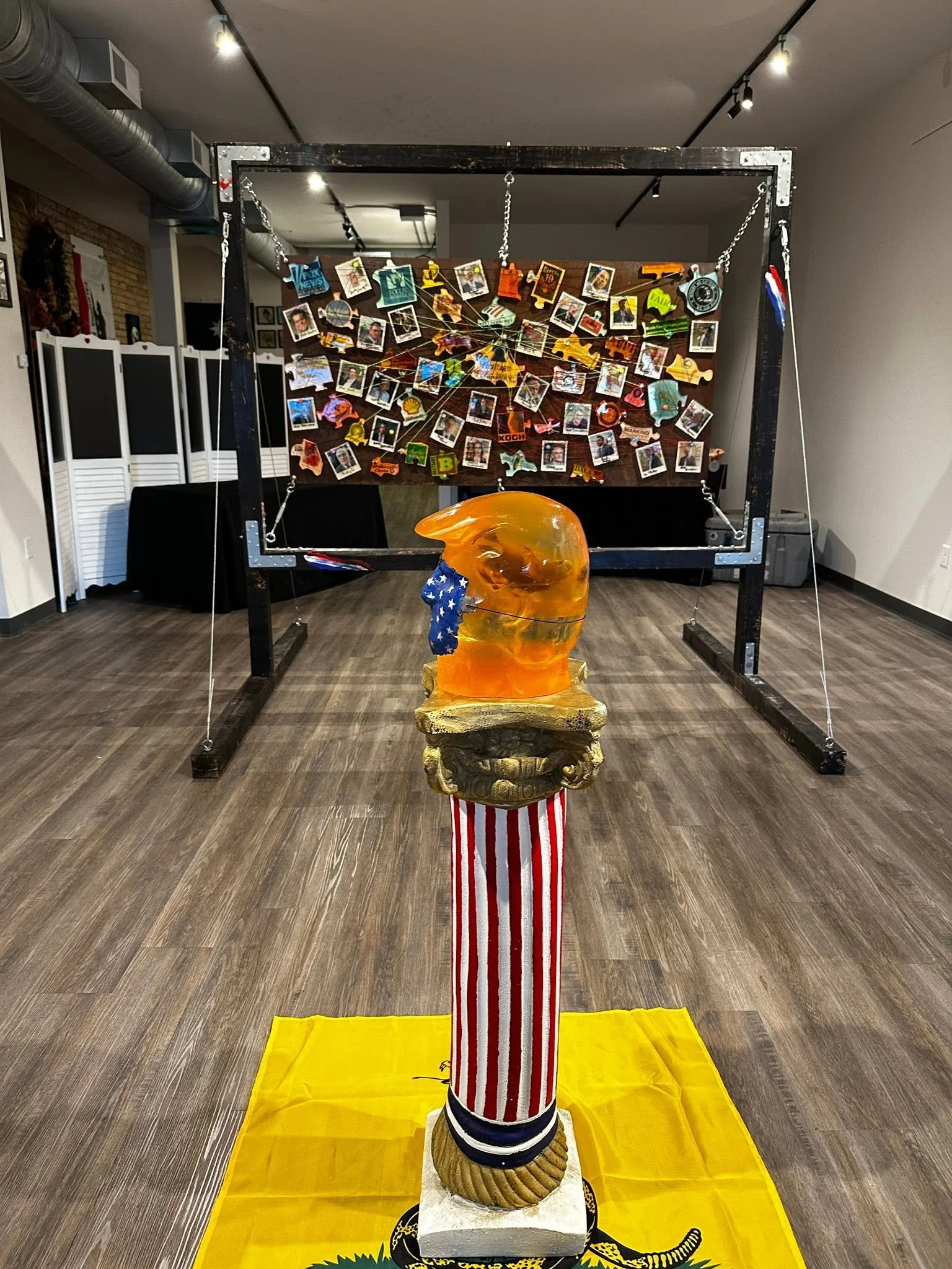 Installation view of Drew Wiggins’s "Thank You for Your Attention to This (Fecal) Matter", featuring a red-and-white Roman column topped with an orange resin head and displayed on a yellow “Don’t Tread on Me” flag.