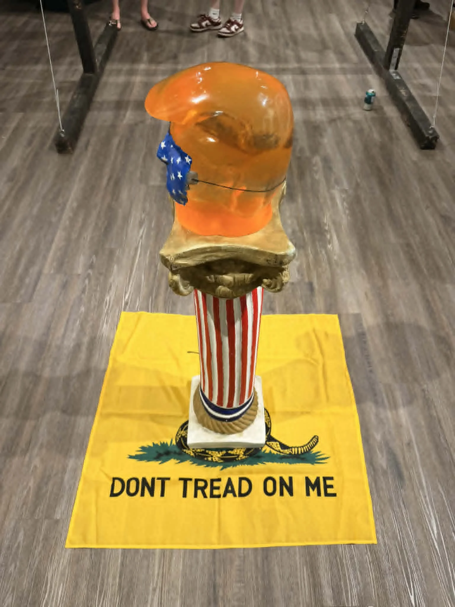 Installation view of Drew Wiggins’s "Thank You for Your Attention to This (Fecal) Matter", featuring a red-and-white Roman column topped with an orange resin head and displayed on a yellow “Don’t Tread on Me” flag.