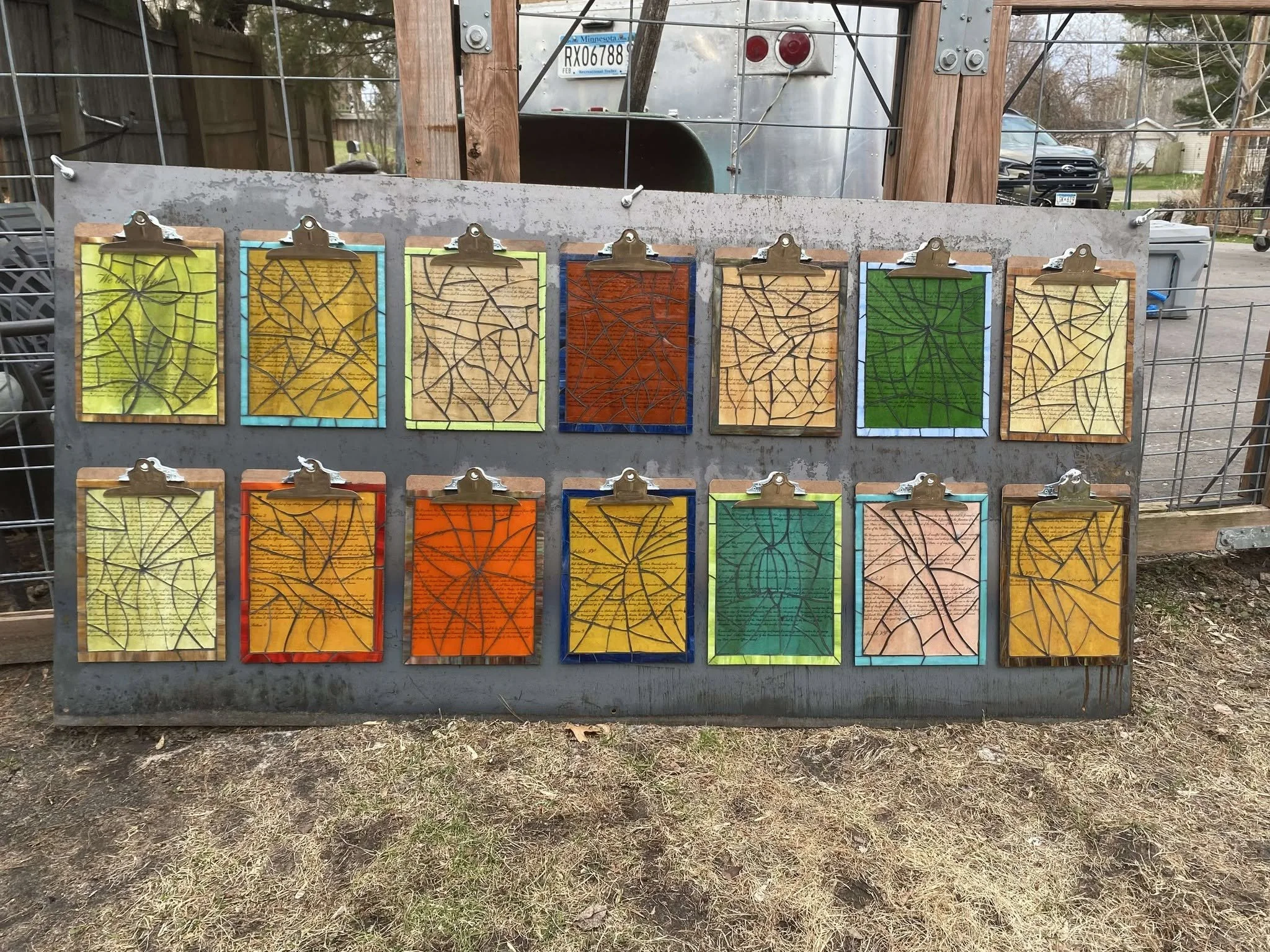 Reverse side of Drew Wiggins’s “Exhibit A: Intentional Malfeasance,” showing fourteen clipboards with brightly colored panels crossed by networks of dark broken-glass lines.
