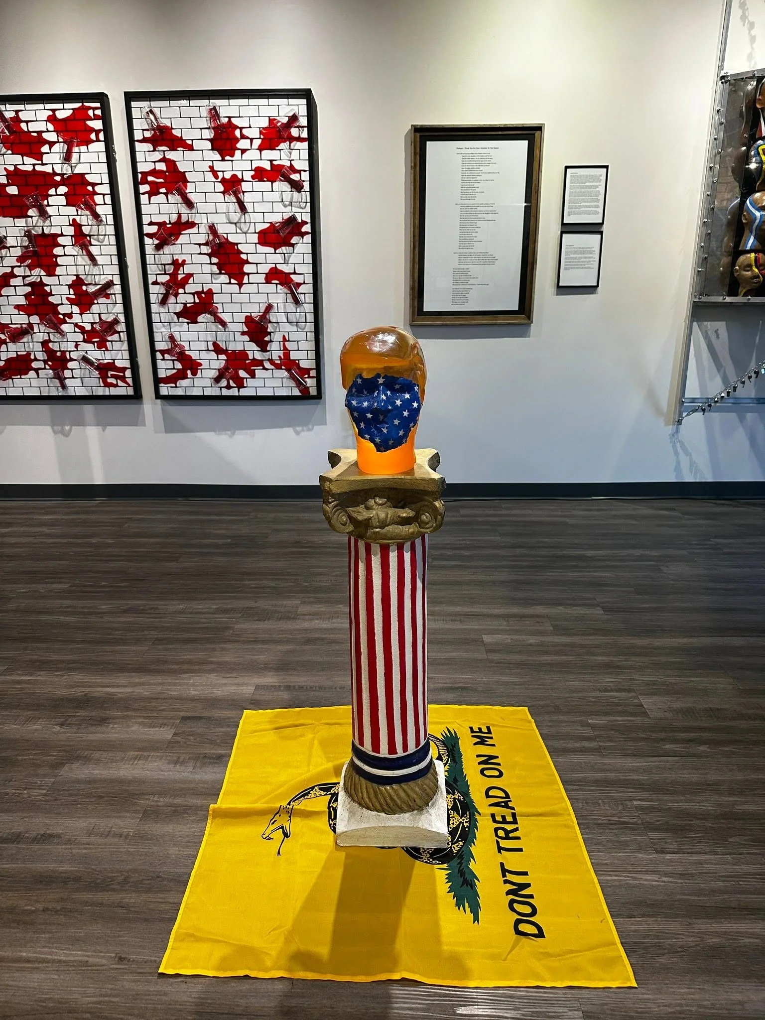Installation view of Drew Wiggins’s "Thank You for Your Attention to This (Fecal) Matter", featuring a red-and-white Roman column topped with an orange resin head and displayed on a yellow “Don’t Tread on Me” flag.