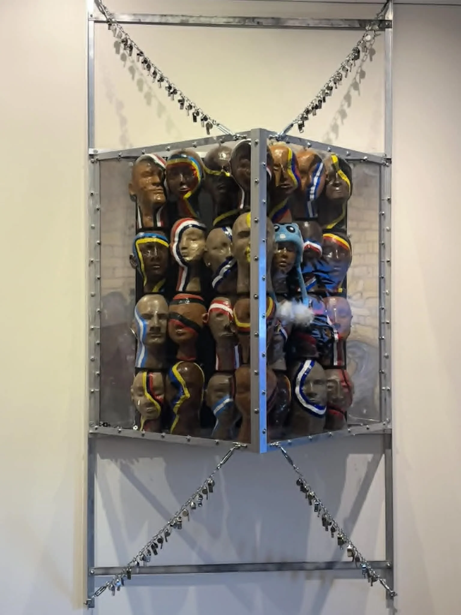 Front view of Drew Wiggins’s “The Cost Per-Capita,” showing a suspended metal enclosure filled with papier-mâché human heads marked by painted bands and stripes.