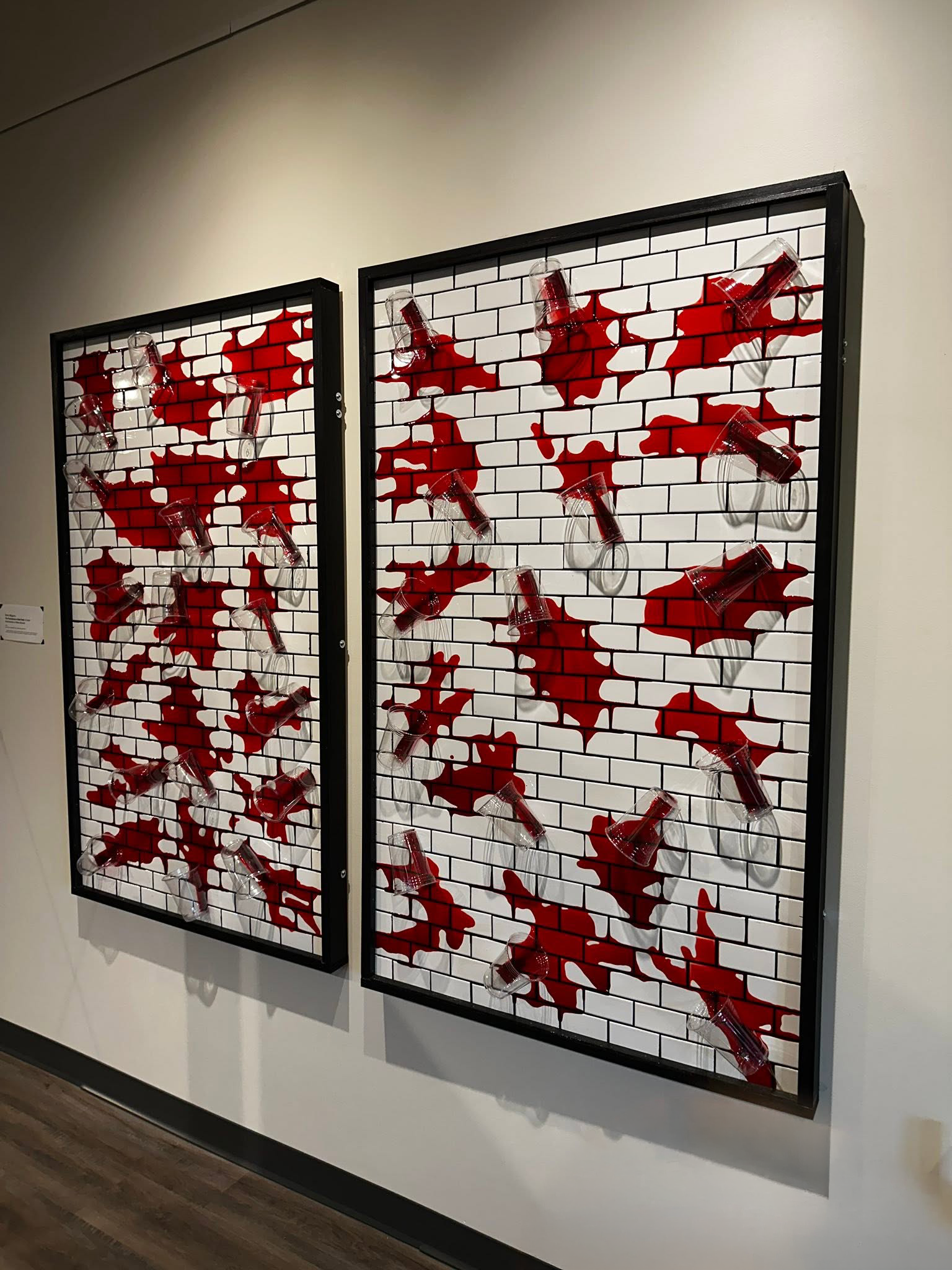 Installation view of Drew Wiggins’s “The Persistence of Bad Faith,” showing two black-framed white subway-tile panels with clear plastic cups and streams of red resin-like liquid spread across the tiled surfaces.