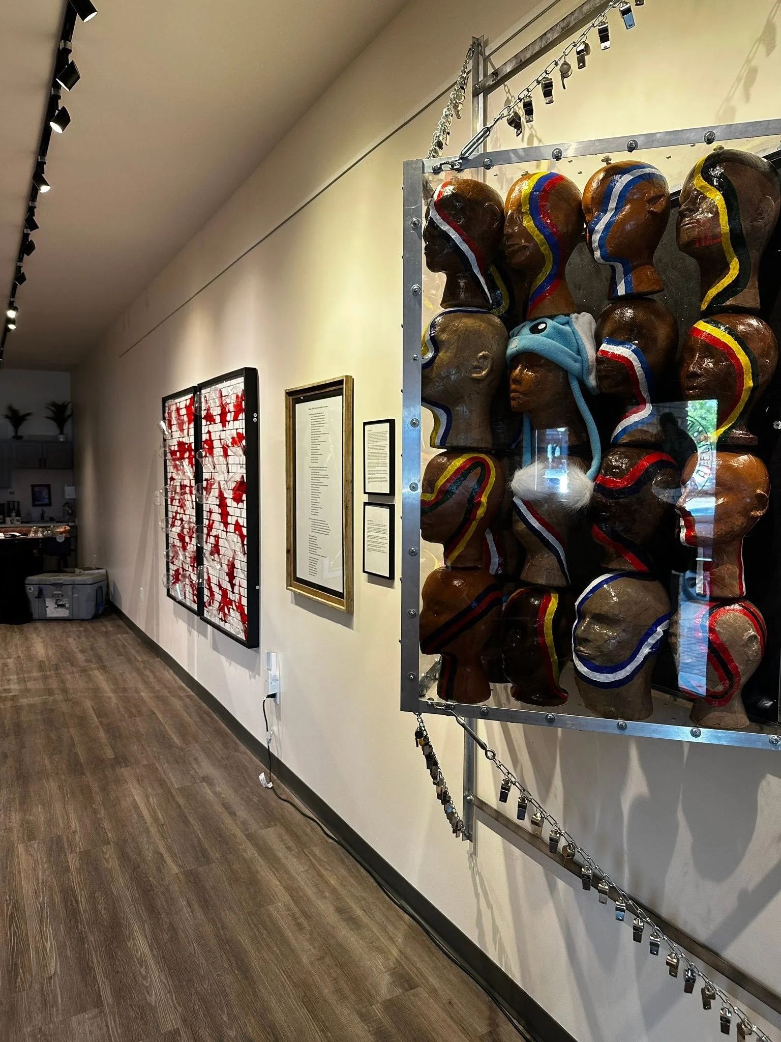 Side installation view of Drew Wiggins’s “The Cost Per-Capita,” showing the clustered papier-mâché heads inside a metal frame, with other exhibition works visible along the gallery wall.