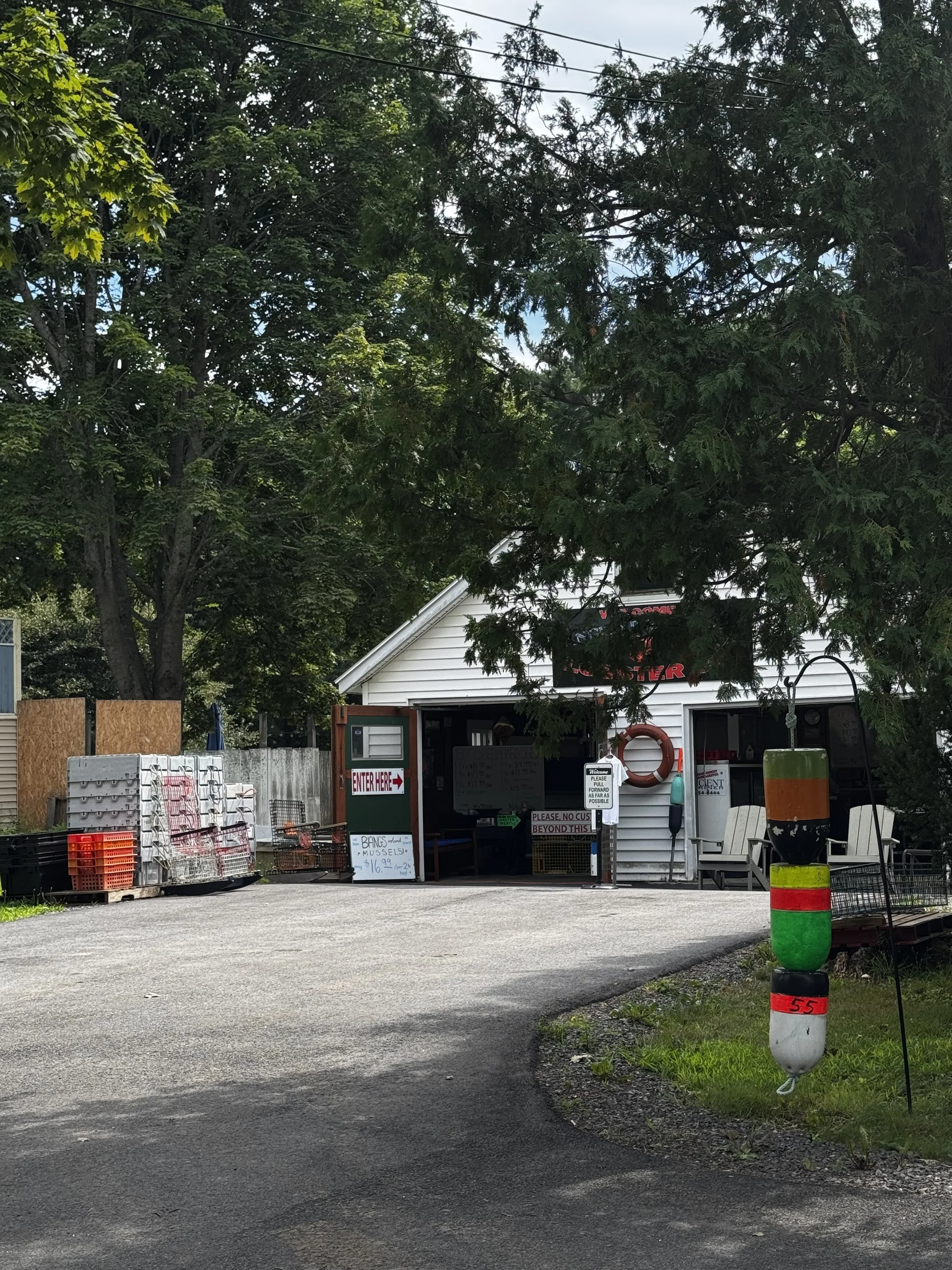 The entrance to a small seafood shop with a white garage exterior and various advertising signs. There are orange and teal buoys as decor out front. Several stacked baskets and crates are on the left. Large trees with green foliage surround the area.