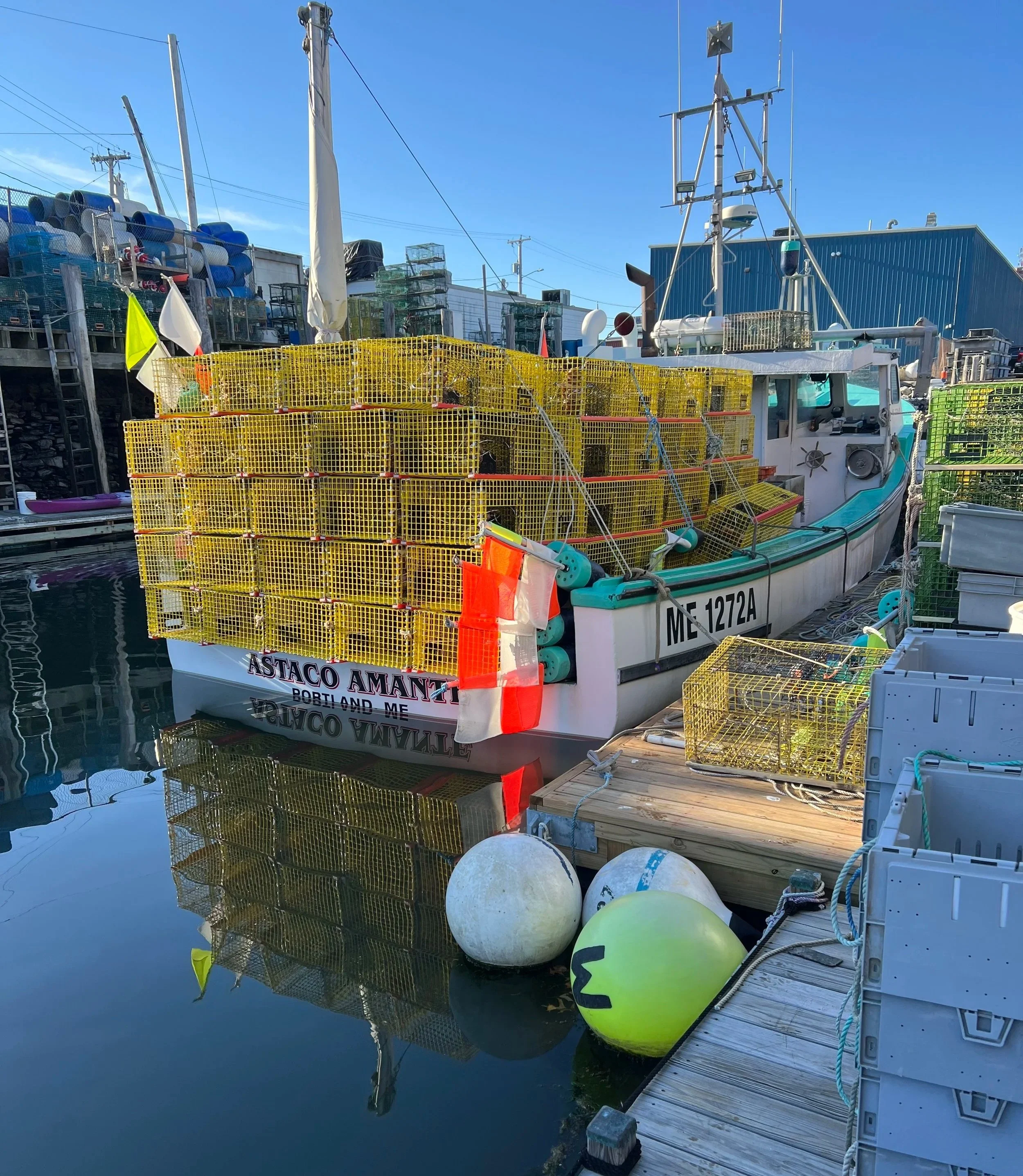 A docked boat with yellow lobster traps, buoys, and fishing equipment, with a blue building and power poles in the background.