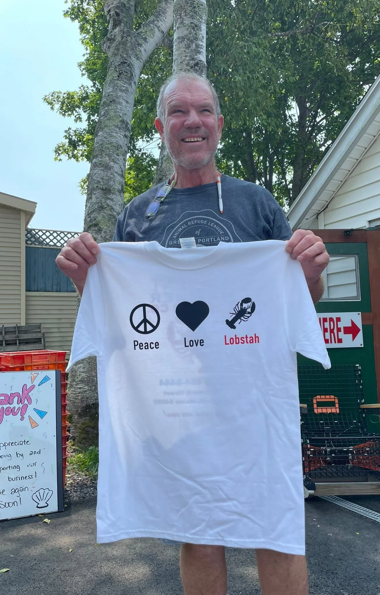 Man smiling outdoors holding a white T-shirt with symbols and words: Peace, Love, Lobstah. Background includes trees, a seafood shop exterior, and a colorful sign that says 'Thank You!'