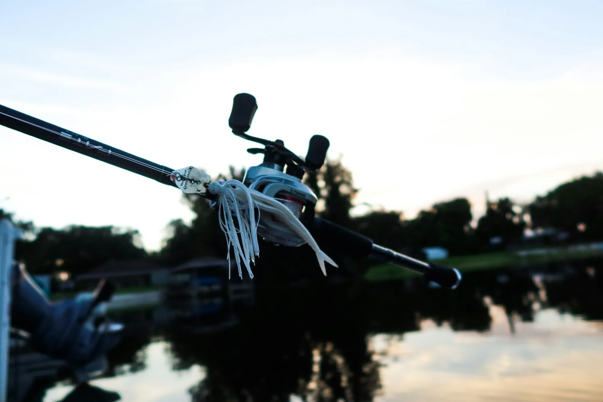 Close-up of a fishing pole with a reel attached, set against a background of a lake or pond with trees and a clear sky during sunset or early evening.