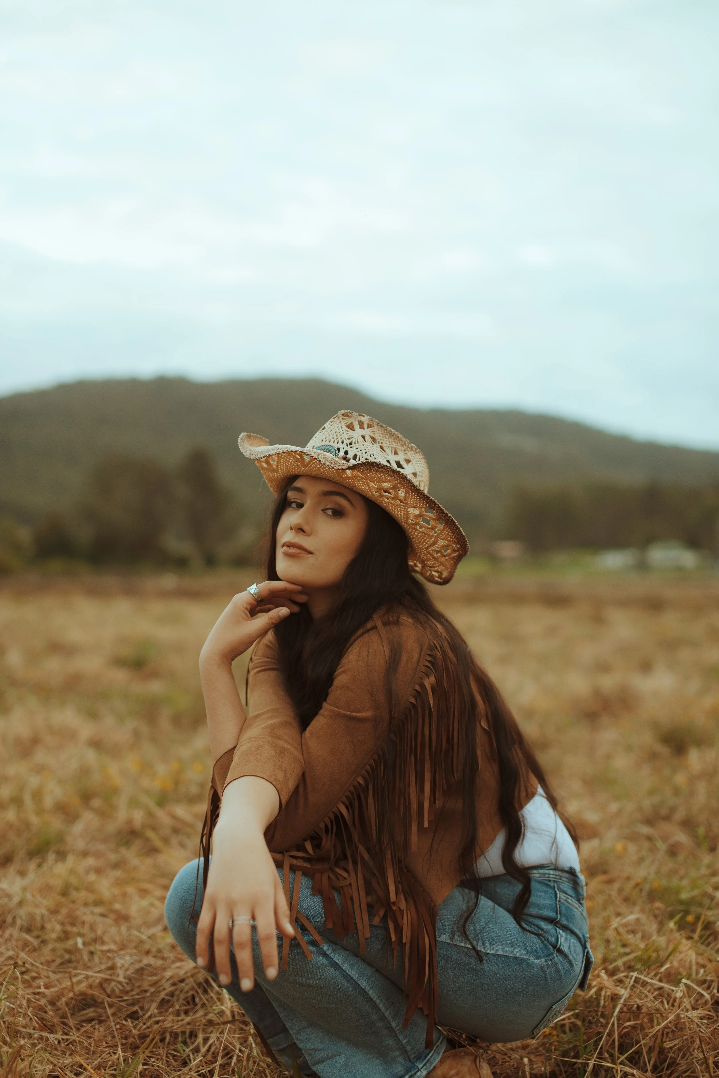 A woman sitting outdoors in a field with mountains in the background, wearing a fringed brown jacket, blue jeans, and a woven cowboy hat.