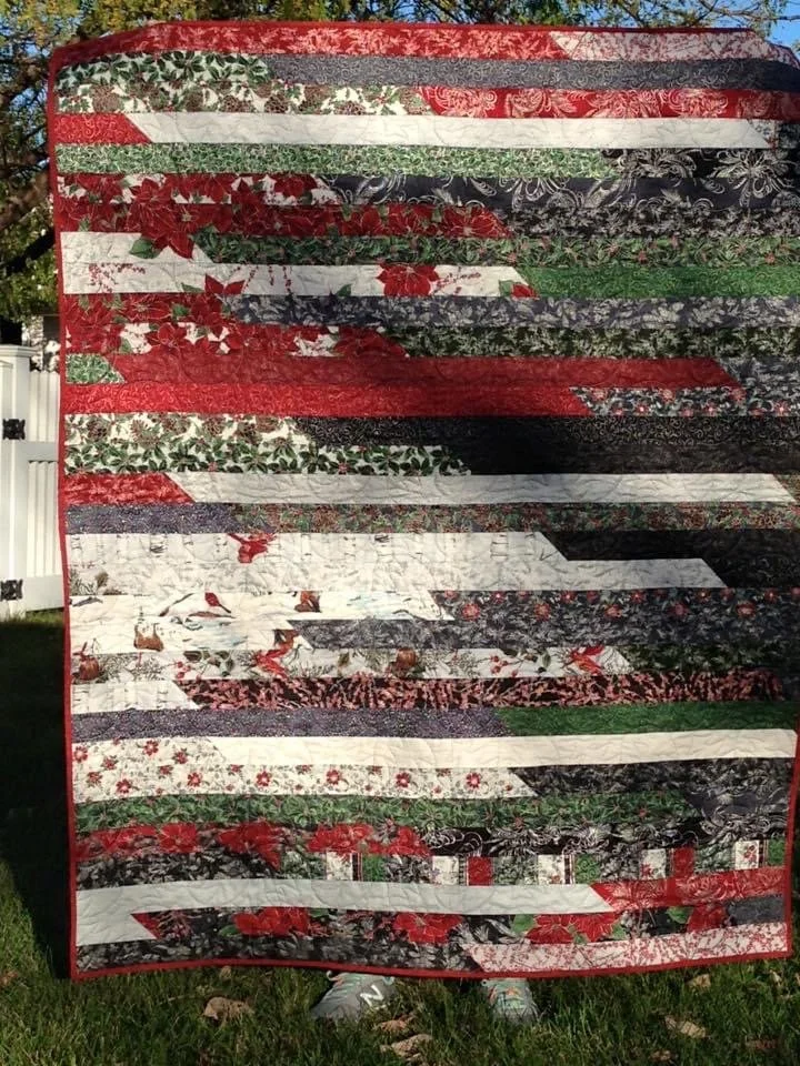 Patchwork quilt with horizontal strips of various holiday-themed fabrics, including poinsettias, holly, and Christmas colors like red, green, black, and white..