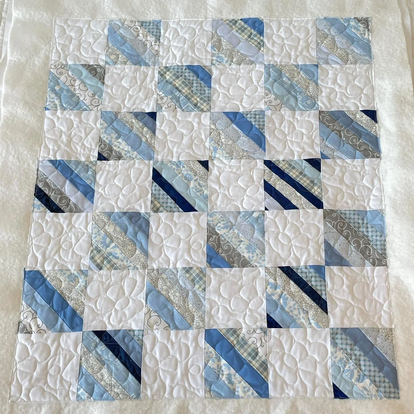 A quilt with a pattern of squares and diagonal strips in shades of blue, gray, and white, with a quilted design of circles and lines.