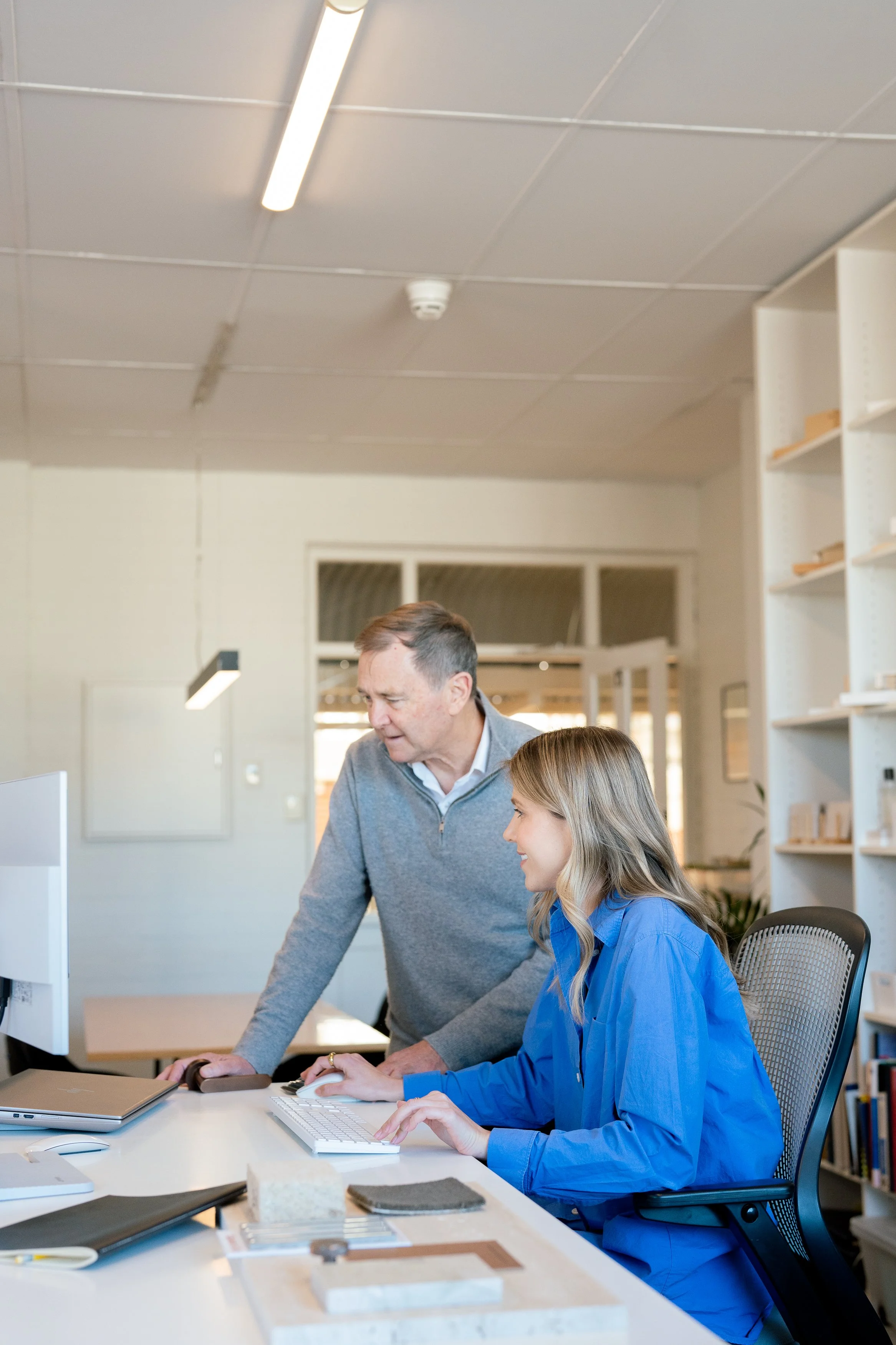 Pia and Paul Hofman working together at a white desk in their bright Cottesloe studio. Pia types at the computer whilst Paul stands next to her giving instruction or explaining something.