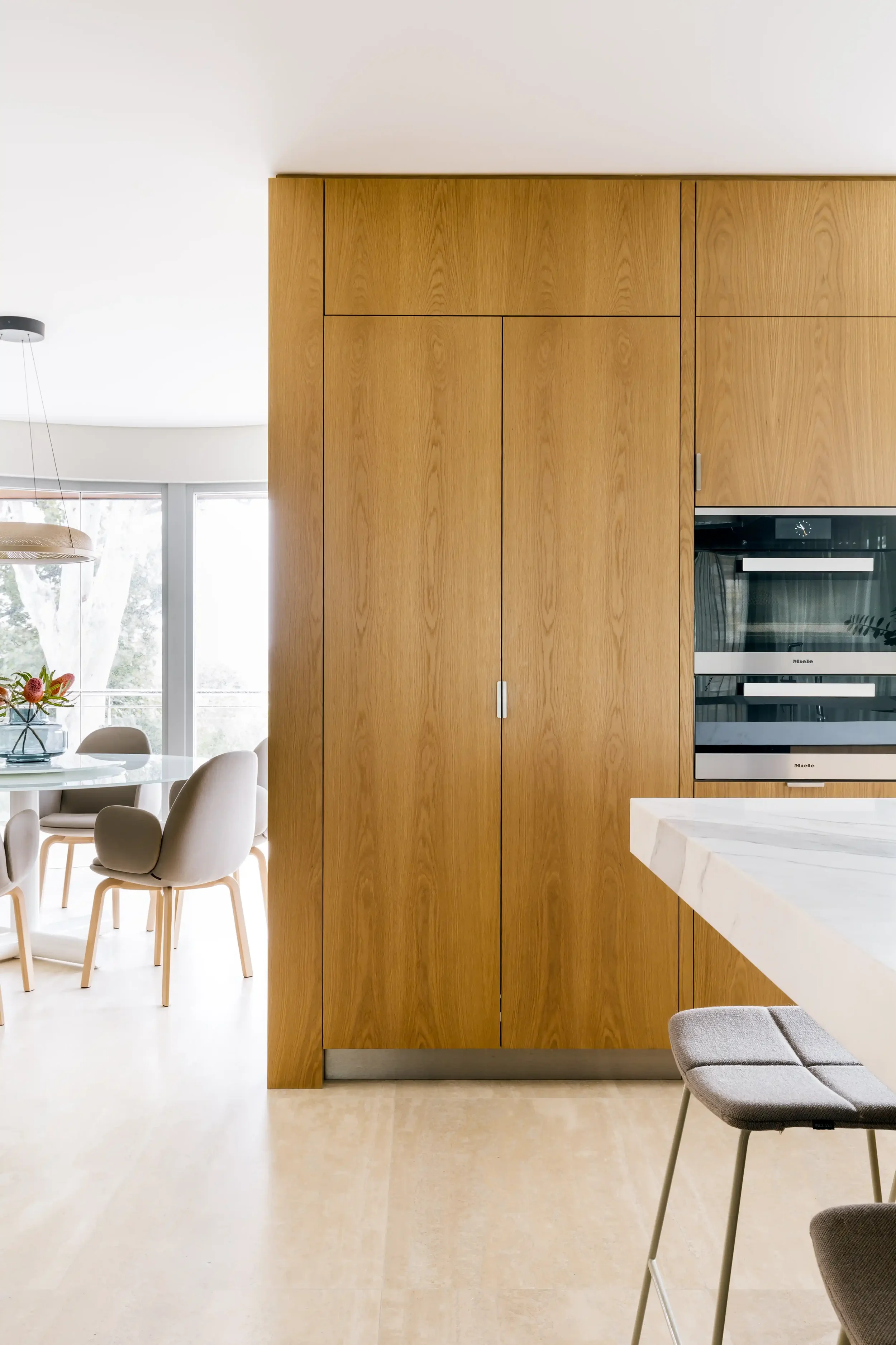 An internal photo of House 40 - Cottesloe. A view of their modern kitchen with warm wooden cabinets, marble countertop, and built-in oven. Adjacent dining area with white chairs around a table, via a window with bright natural light.