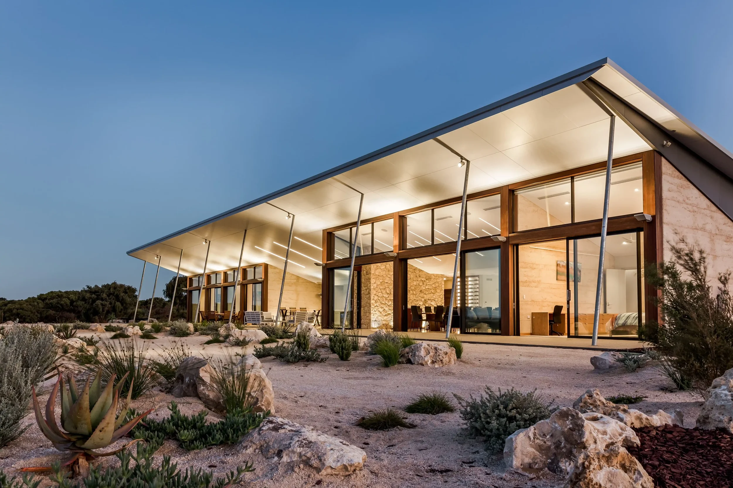 An external photo of House 214 - Yallingup. This home has floor to ceiling glass windows and wooden accents, illuminated from within, set in South of WA landscape with rocks and succulent landscaping, during dusk or early evening.