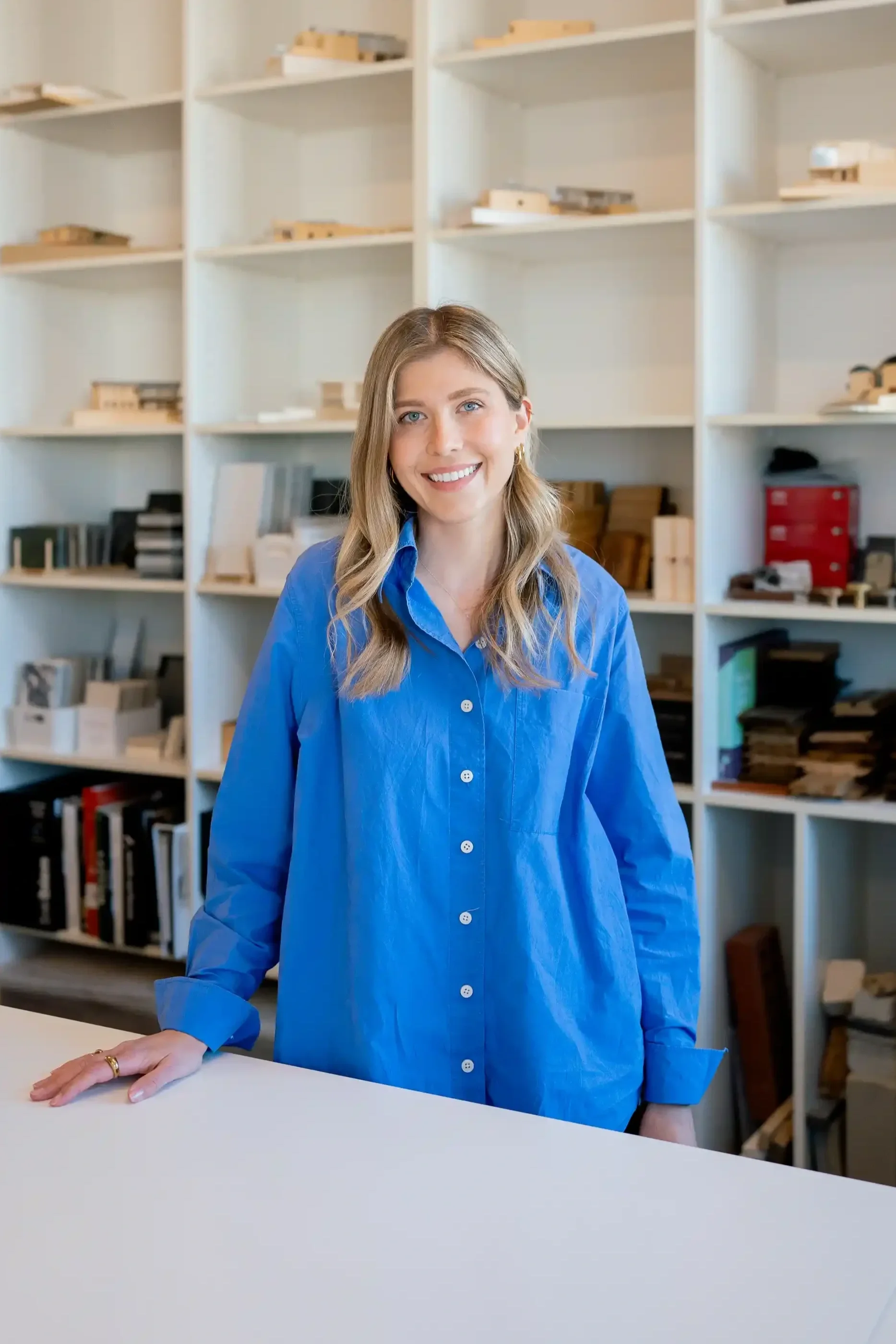 Pia Hofman, smiling with long blonde hair wearing a blue button-up shirt, standing in front of white shelves filled with various wooden models and office supplies, resting her hand on a white table in their modern Cottesloe studio.