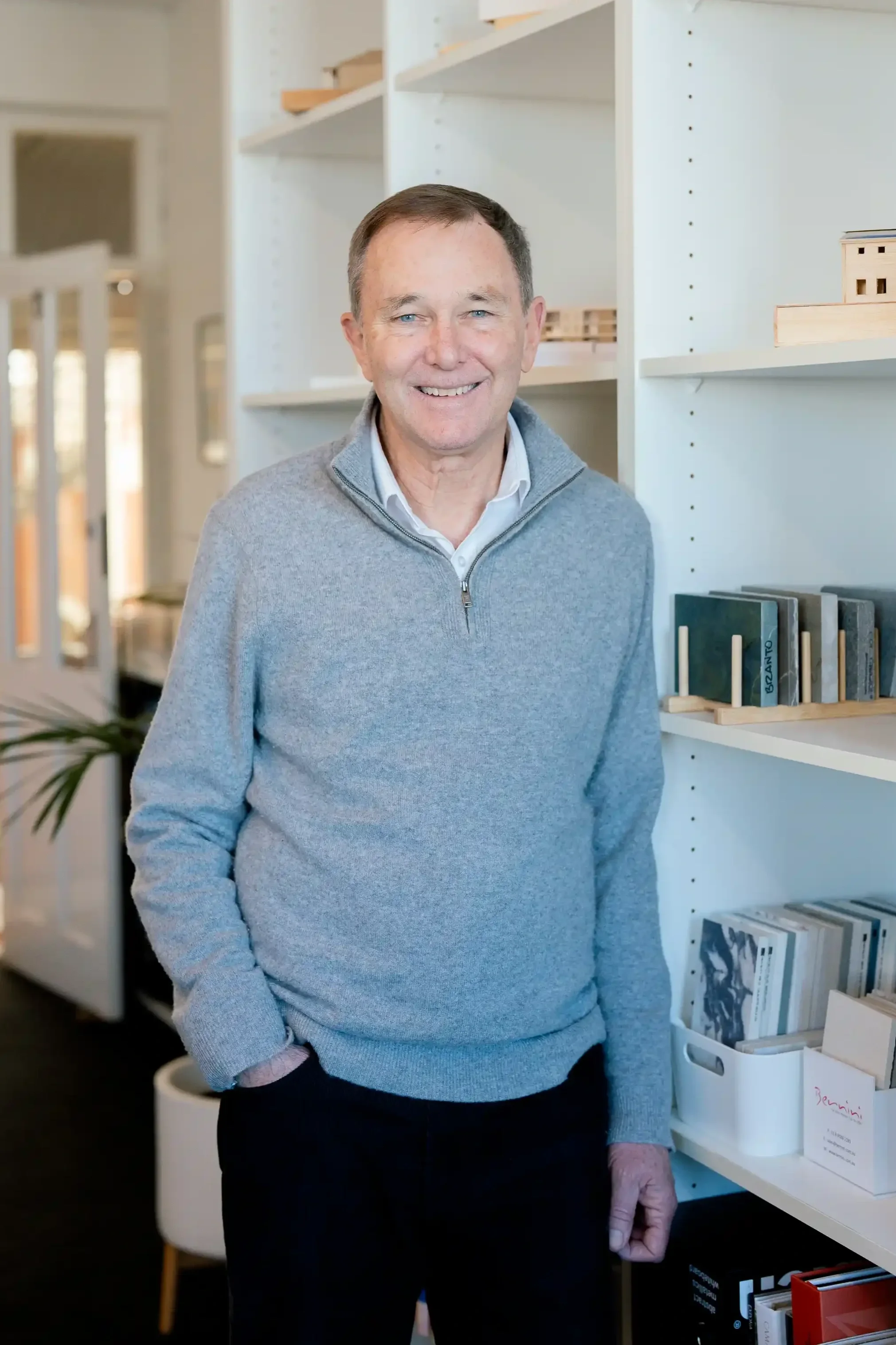 Paul Hofman, with short brown hair and blue eyes, smiling, wearing a light grey quarter-zip sweater over a collared shirt, standing in front of white bookshelves filled with books and wooden model structures in their modern Cottesloe studio.