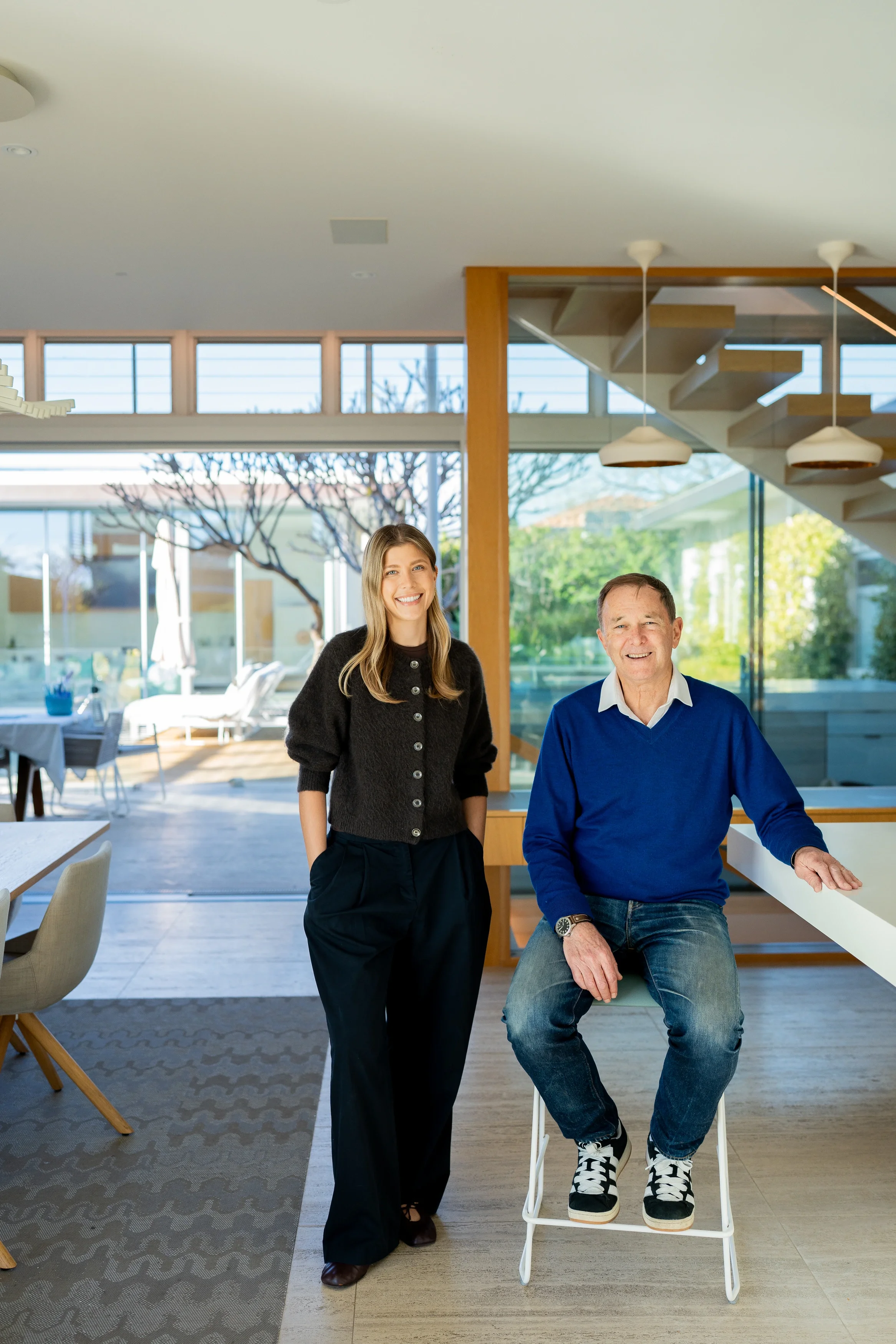Pia and Paul Hofman sitting in House 40 - City Beach, a modern, bright home with large windows, trees outside, and contemporary decor.