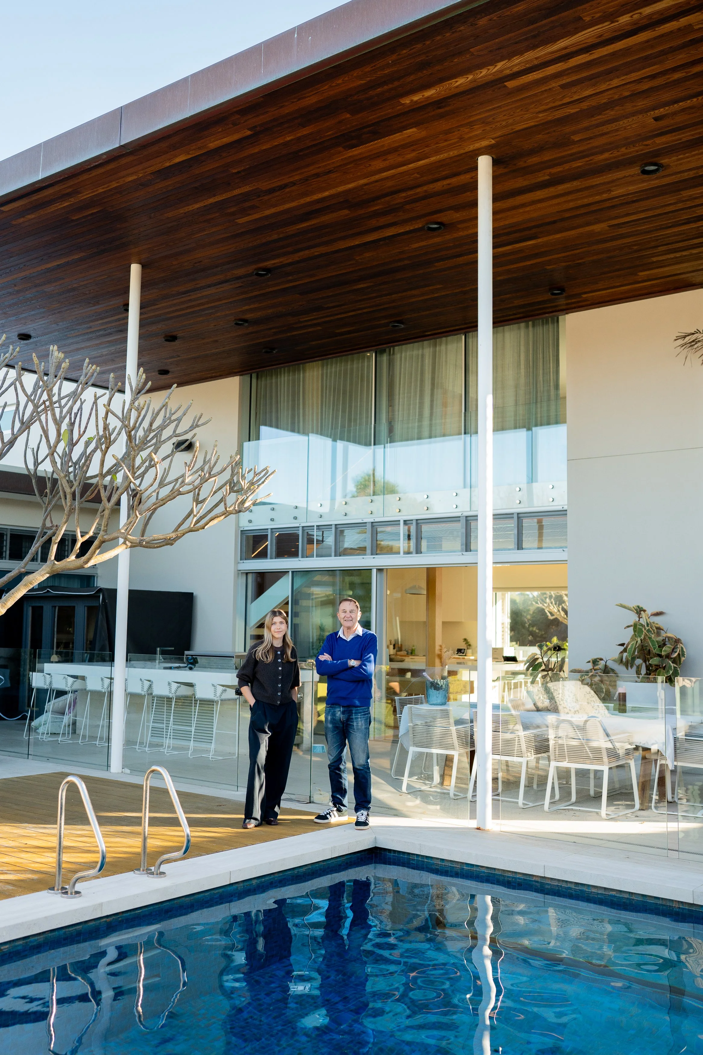 Paul and Pia Hofman standing by the swimming pool in front of a House 40 - City Beach. Large glass doors stand behind them, a wooden overhang above, styled with outdoor chairs and tables.