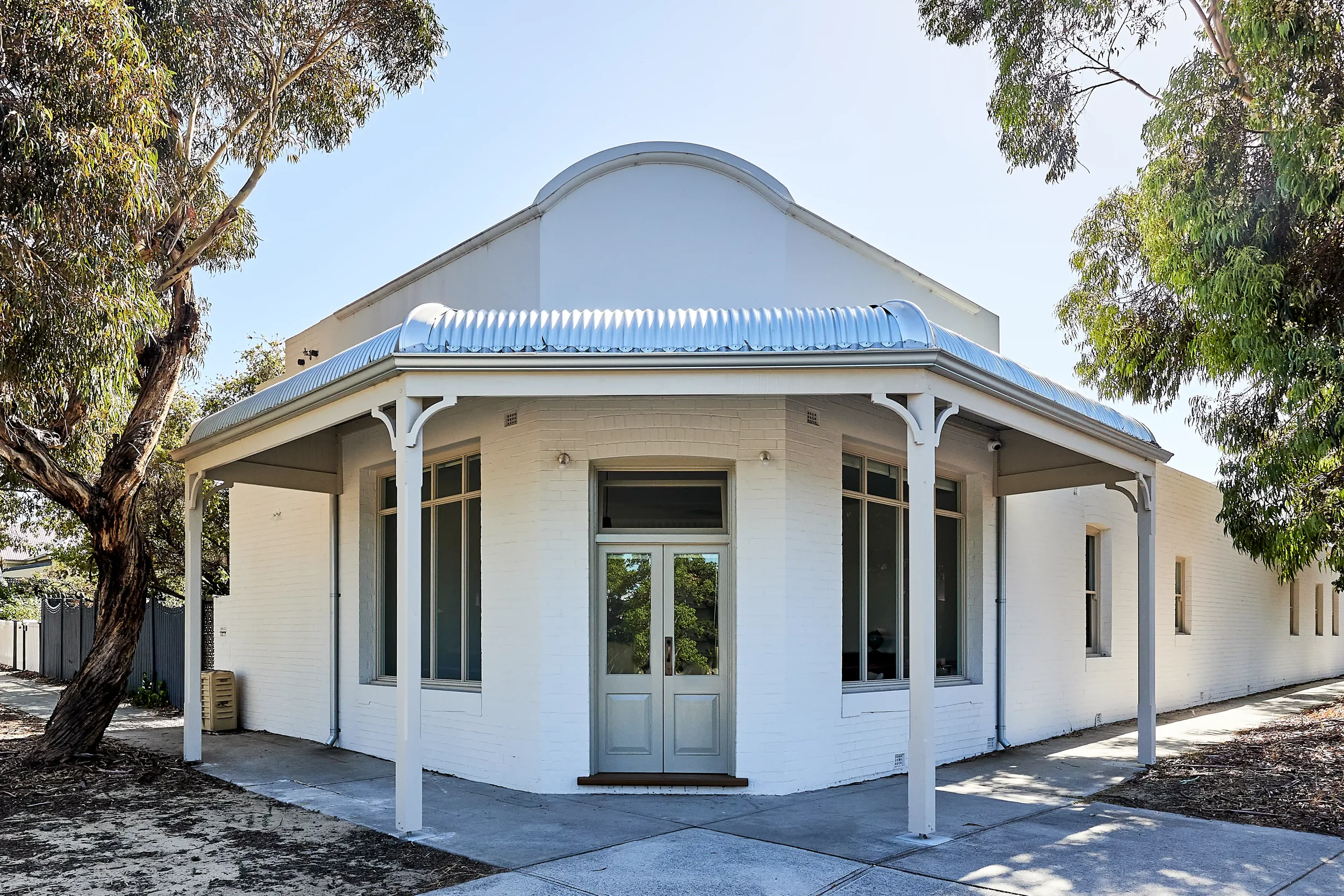 An external photo of The Old Corner Stone Project. A heritage style white home with a rounded roof and large windows, surrounded by trees and a sidewalk, under a clear blue sky. This was a corner store that was converted into a home.