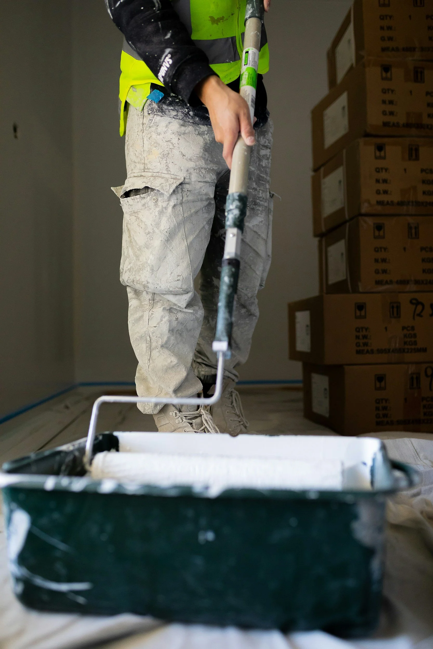 A construction worker in dusty white pants and a neon yellow safety vest is holding a paint roller. The worker's face and upper body are not visible, and they are standing near stacks of cardboard boxes inside a room under construction.