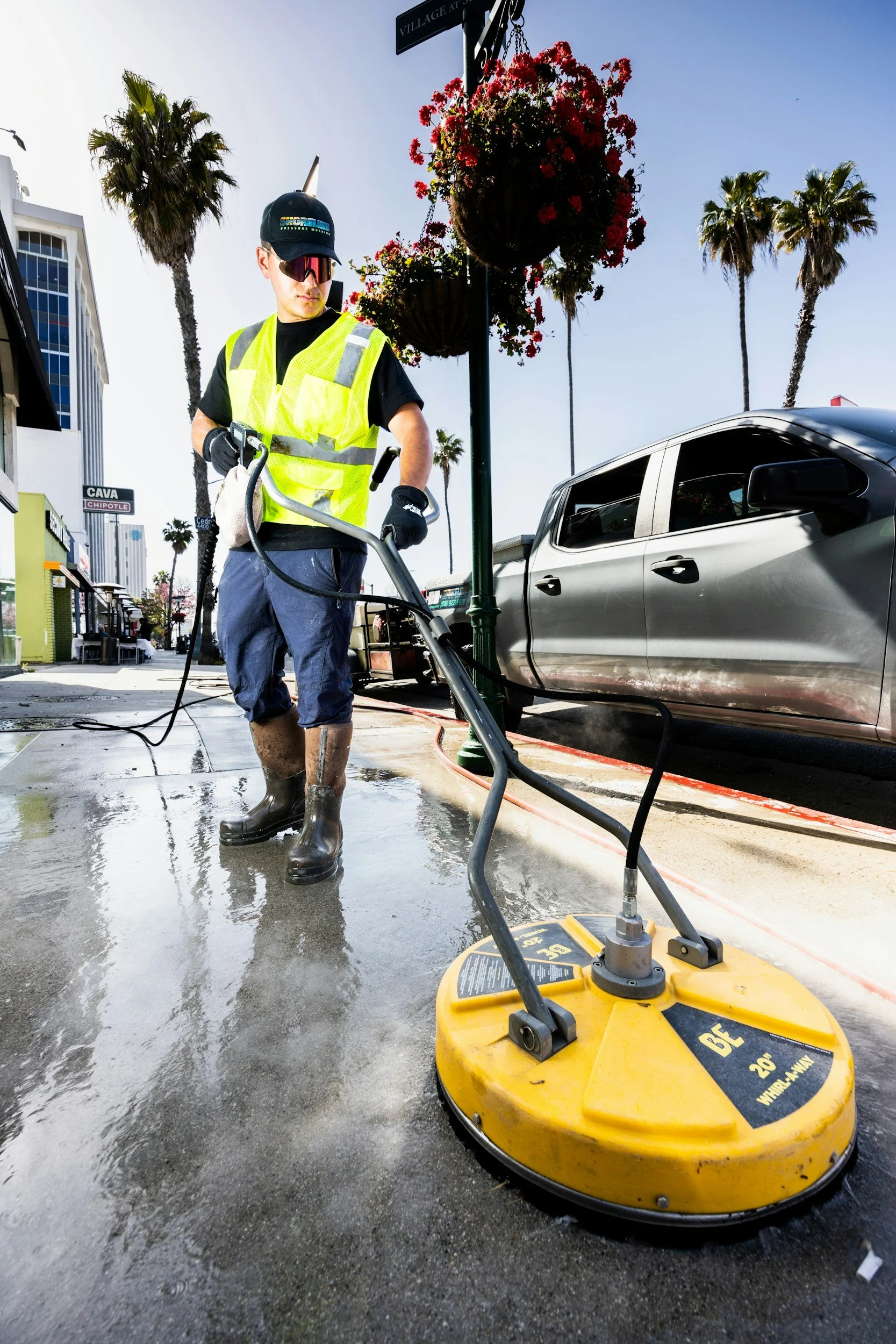 A man wearing a yellow safety vest, black shirt, and brown rubber boots is operating a yellow floor buffer machine on a wet sidewalk with steam or water spraying. He is wearing glasses and a black cap. There are palm trees, a silver car, and storefronts, including a Chipotle restaurant, in the background on a sunny day.