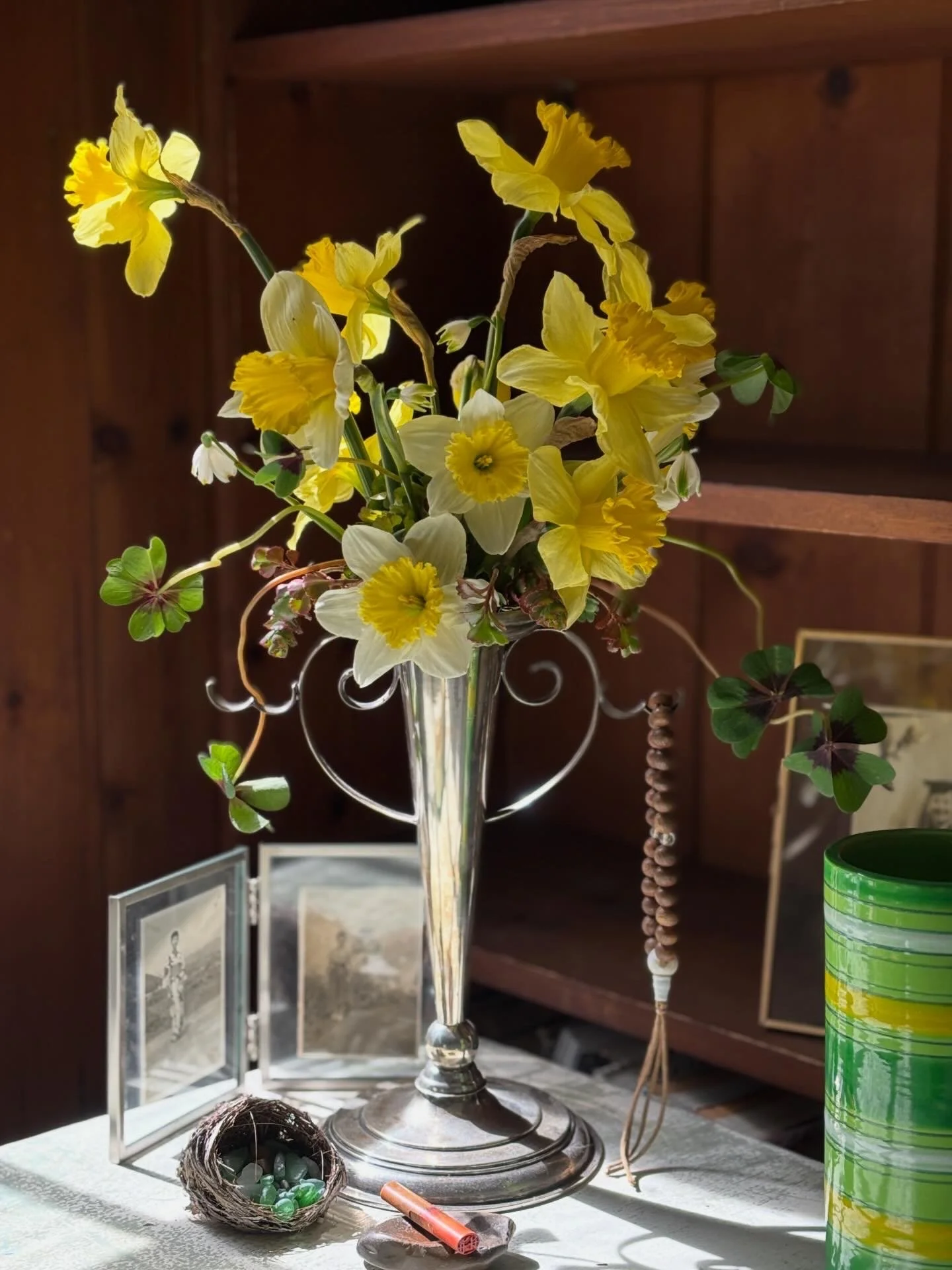 A tall clear glass vase holding a bouquet of bright yellow daffodils and some small white flowers with dark centers. There are several clovers with green leaves and three leaflets, and some green marbles in a small nest at the base of the vase. A small carved wooden object and a red pen are also on the table. In the background, there is a wooden cabinet with framed photographs or artwork.