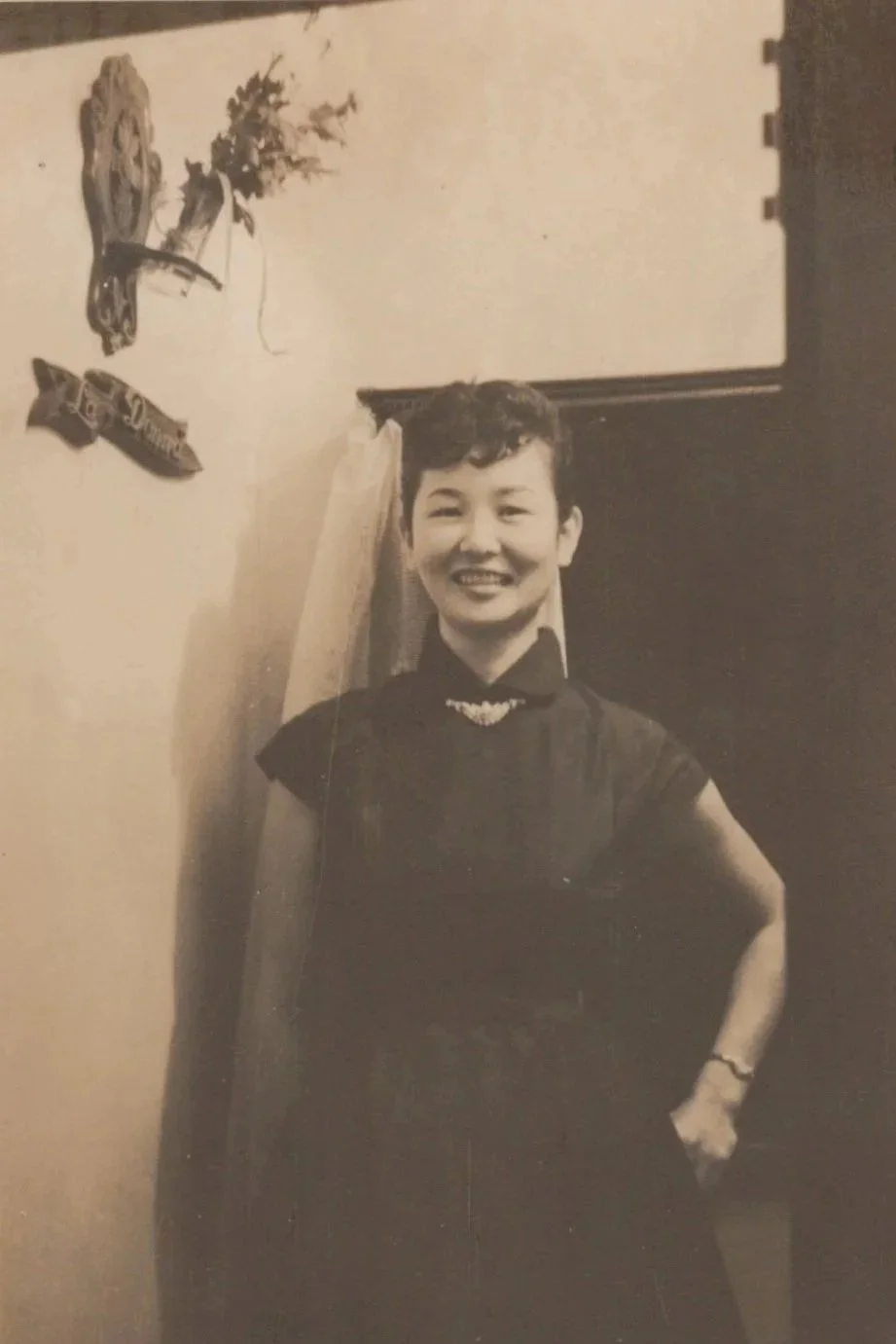 A vintage black and white sepia toned image of a young Japanese woman in a black dress with a high collar, with black hair posing in front of a doorway next to a sign reading "La Donna" and a  hanging floral arrangement.