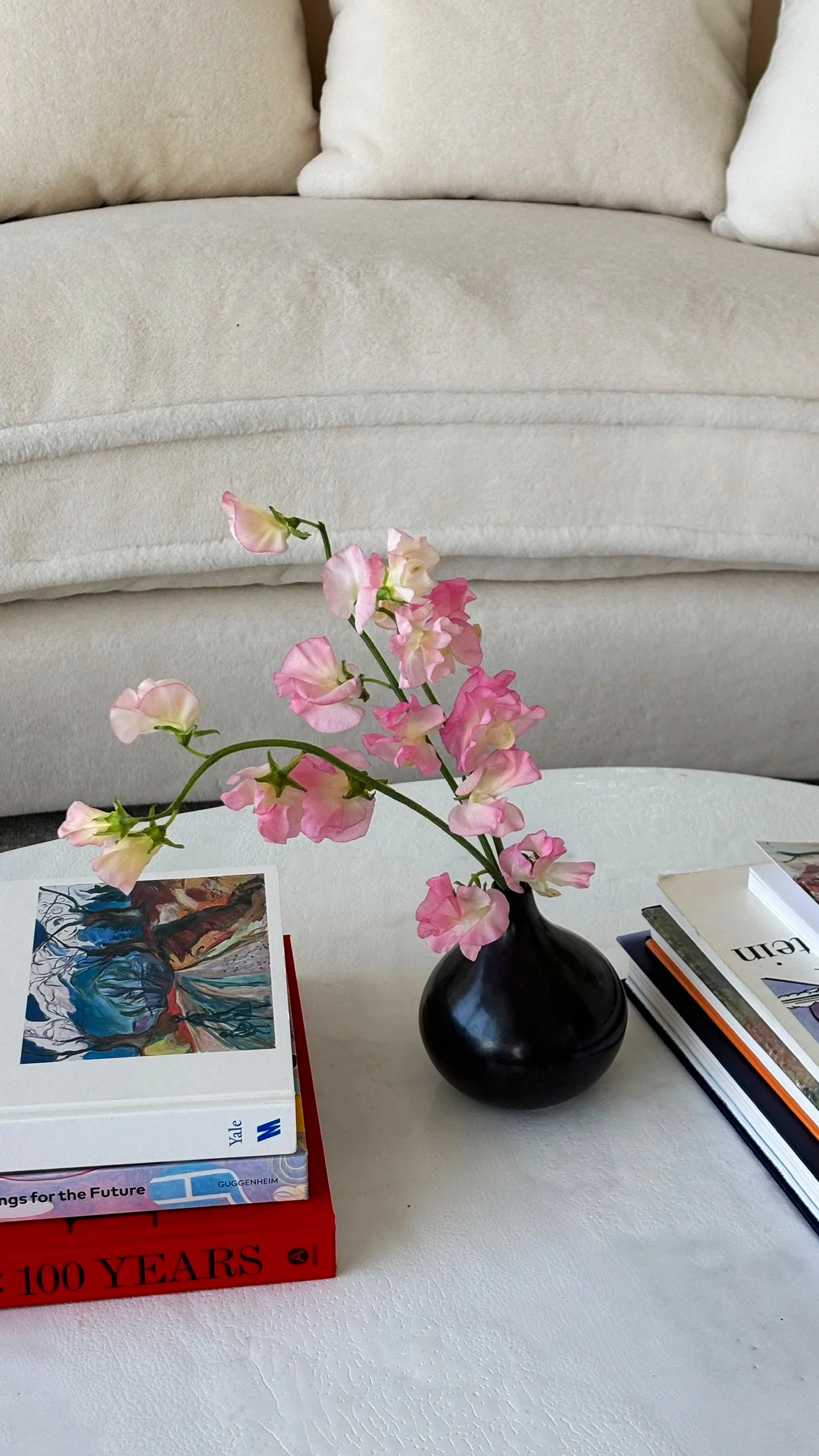 A black vase with pink and white flowers on a white table, with books and magazines around it, in a room with a beige sofa in the background.
