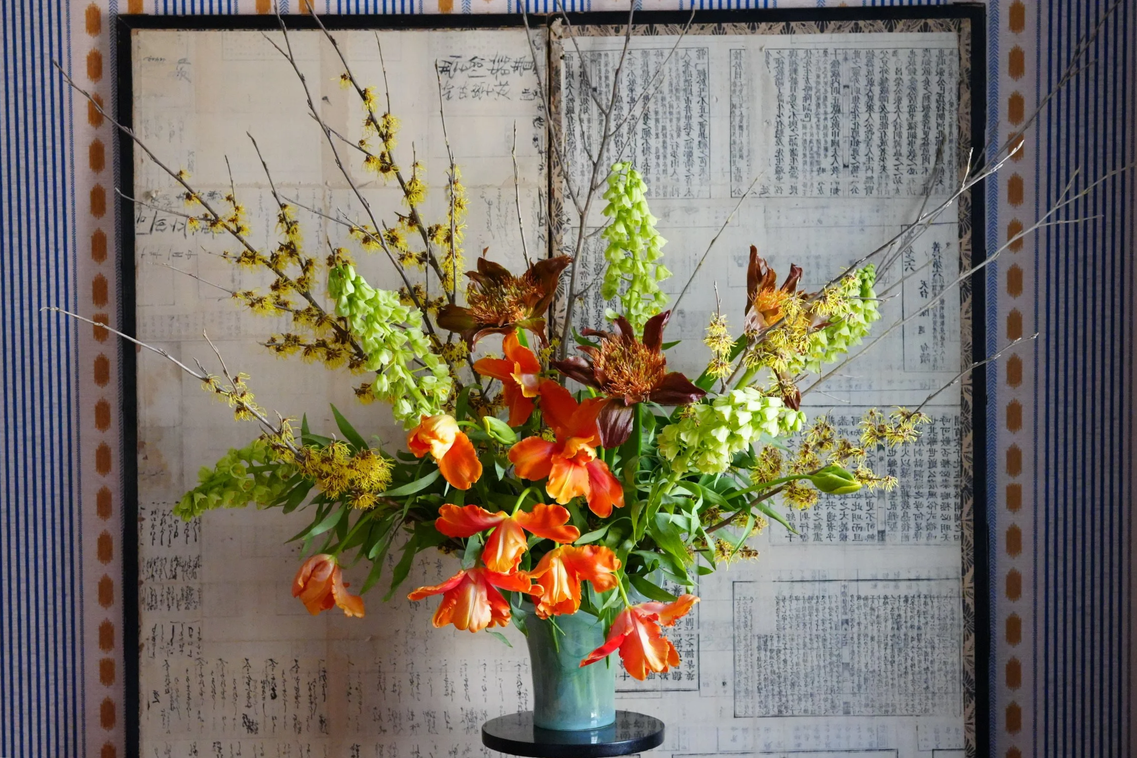 A colorful floral arrangement with orange, yellow, and green flowers in a blue and green vase, set against a background of patterned fabric and Asian calligraphy.
