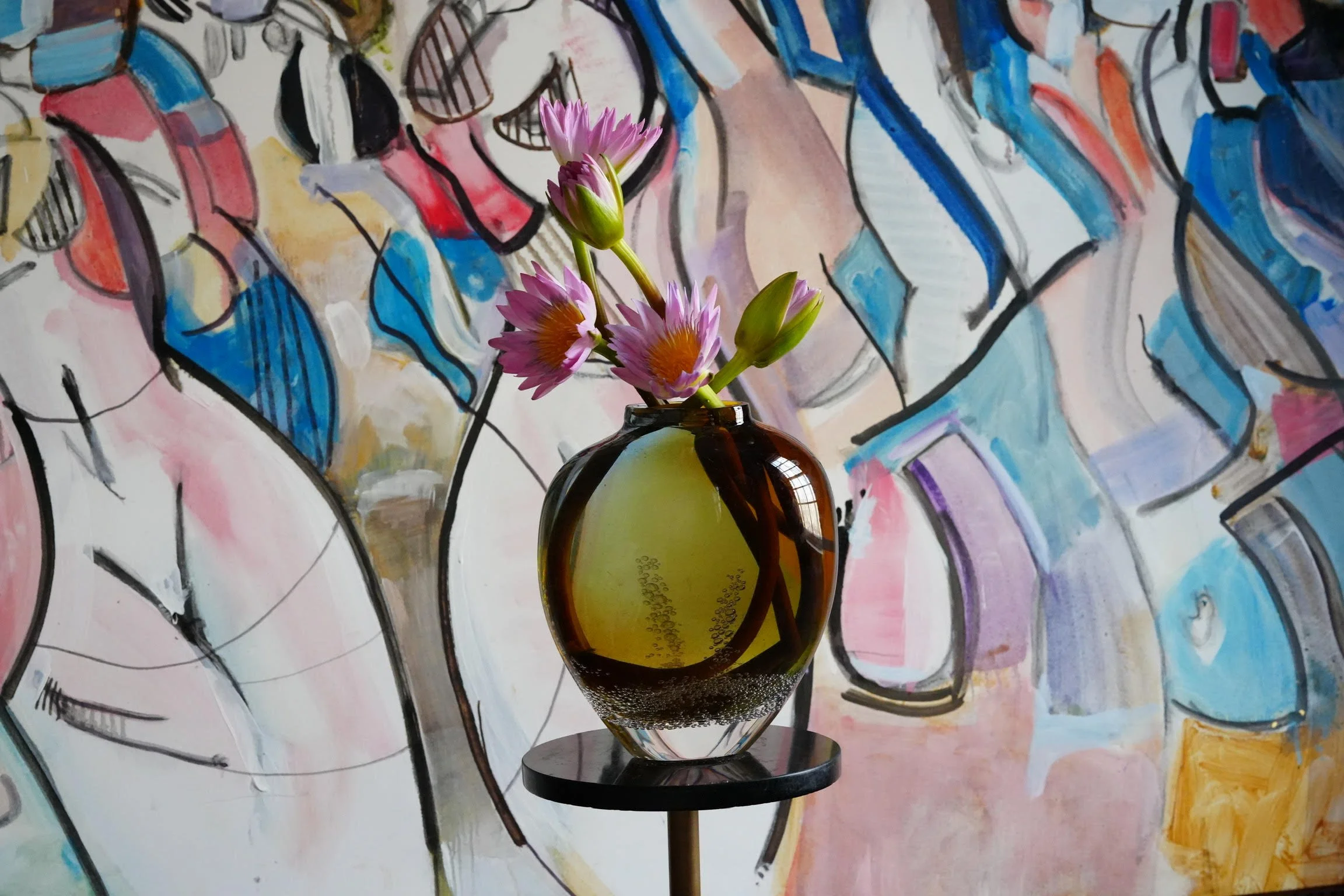 A small bouquet of pink and purple flowers in a brown glass vase placed on a black stand, with an abstract colorful painting in the background.