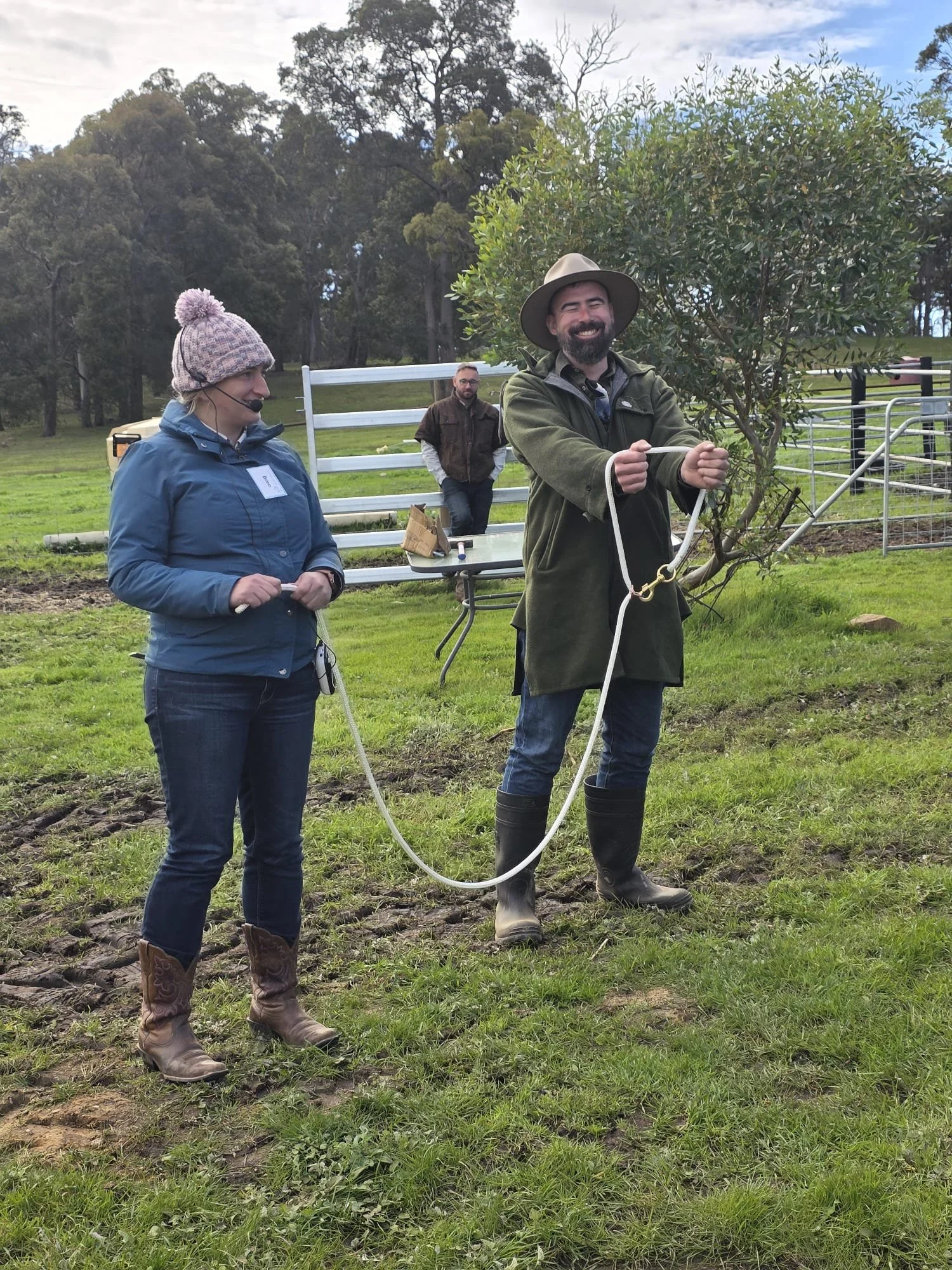 Three people in an outdoor setting with trees and grass. Two are in the foreground; one woman with a microphone headset and a blue jacket, and a man with a wide-brimmed hat and green jacket holding a rope. A third man is in the background near a white fence, wearing a brown jacket and standing by a table with some items on it.