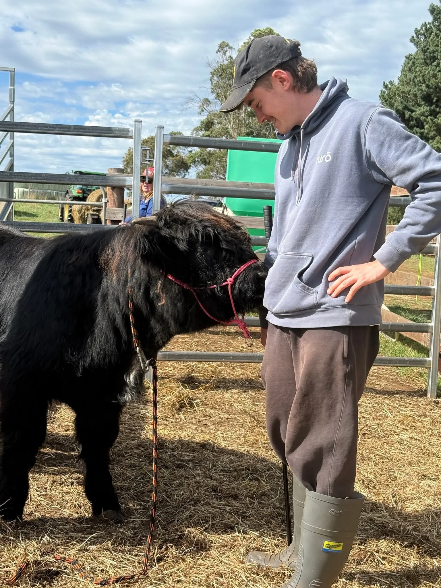 A young man in gray waterproof boots and a gray hoodie stands in a rural farm setting, holding a black and white calf by a pink rope, with a young girl wearing sunglasses and a pink hat in the background near a metal fence and green farm equipment under a cloudy sky.